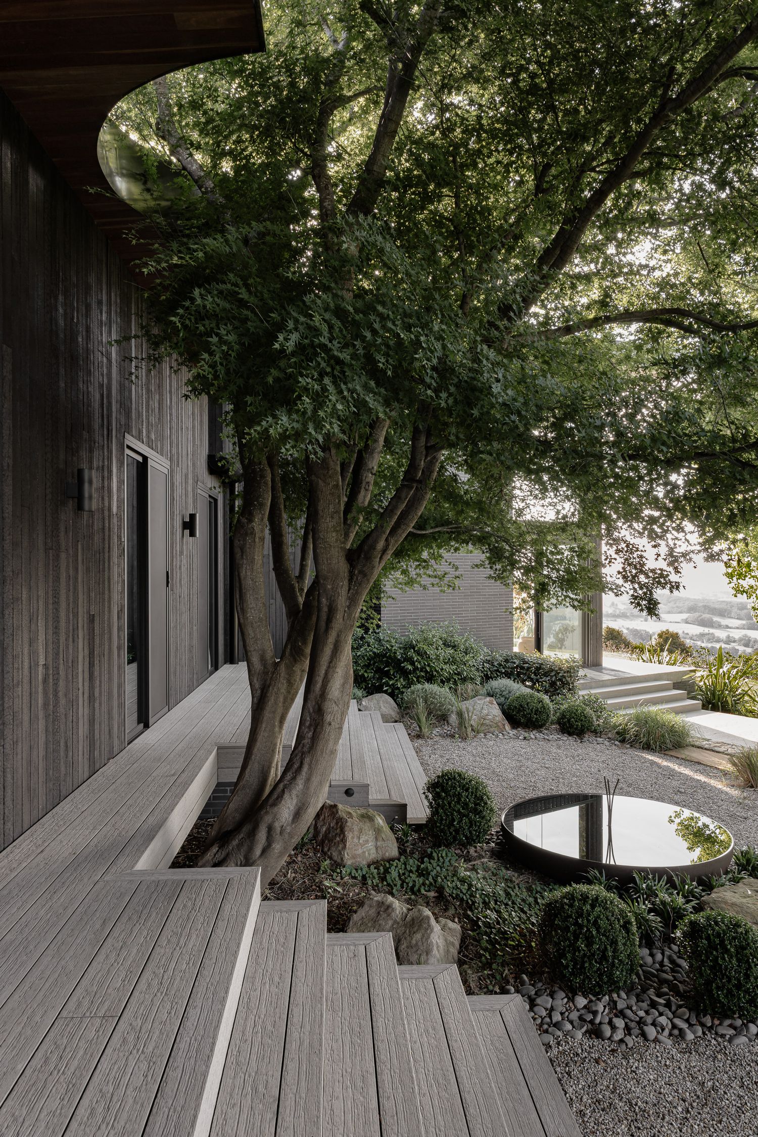 Modern wooden deck beside a zen garden with manicured shrubs, gravel, and a large tree framing a contemporary dark-clad home.