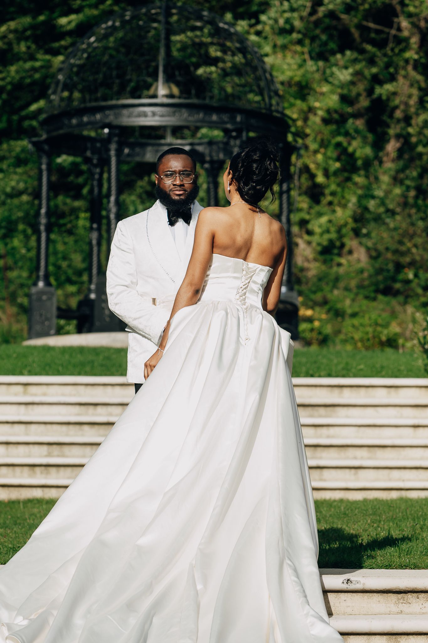 A bride and groom in elegant white attire share a romantic moment in a lush garden with a decorative gazebo.