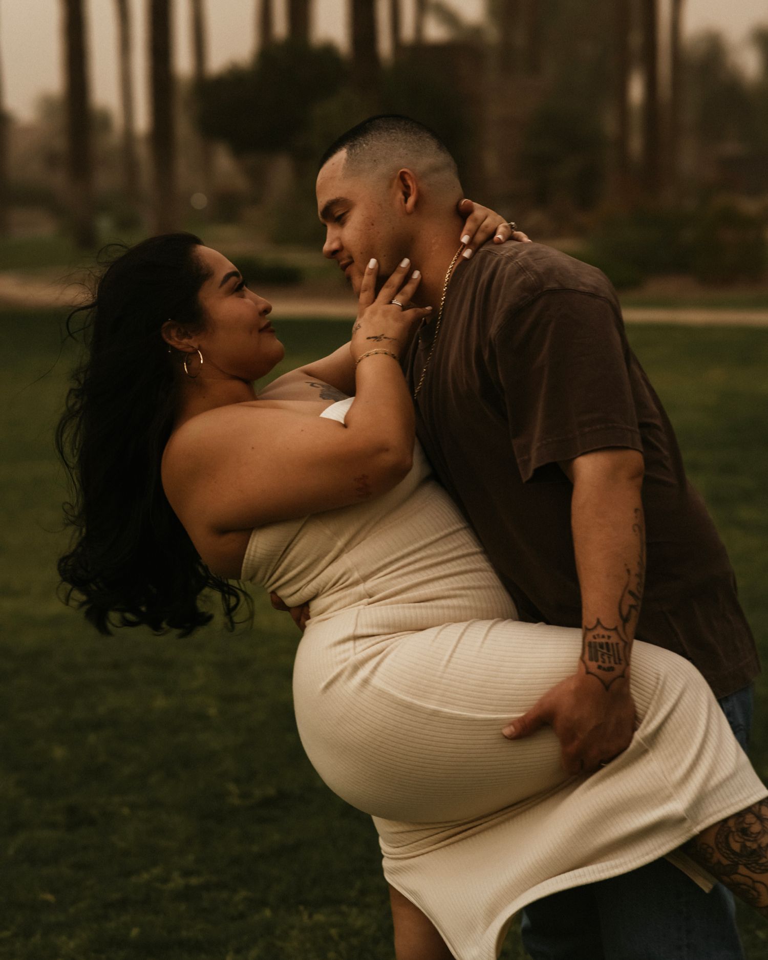 A man lifts and embraces a woman in a cream dress outdoors, sharing a romantic moment in a lush green park.