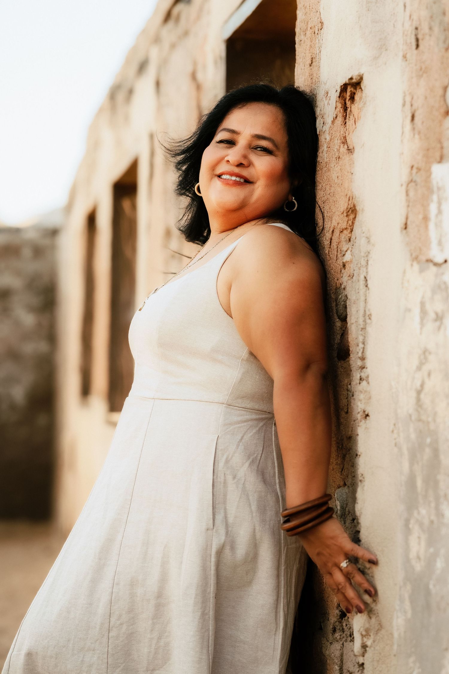 A person in a white summer dress poses against a rustic stone wall in warm sunlight.