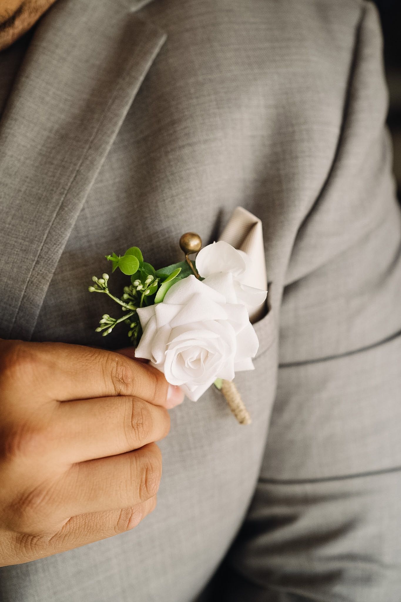 Hand pinning a white rose boutonniere with greenery onto a gray suit lapel for a wedding ceremony.