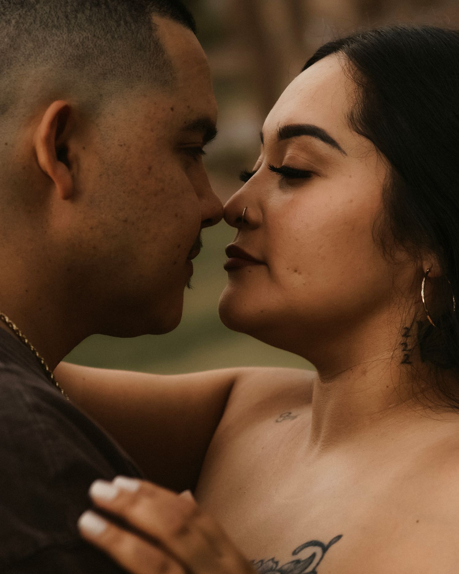 A couple shares an intimate almost-kiss moment, with the woman showing hoop earrings and a shoulder tattoo.