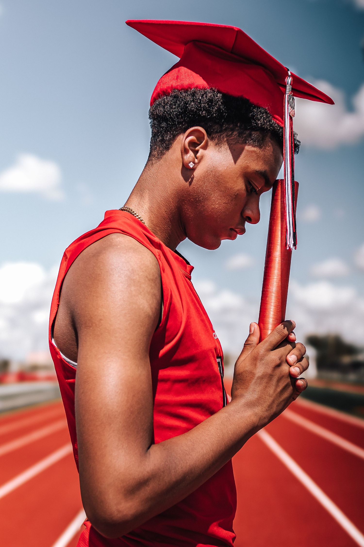 Graduate in red cap and gown stands reflectively on running track.