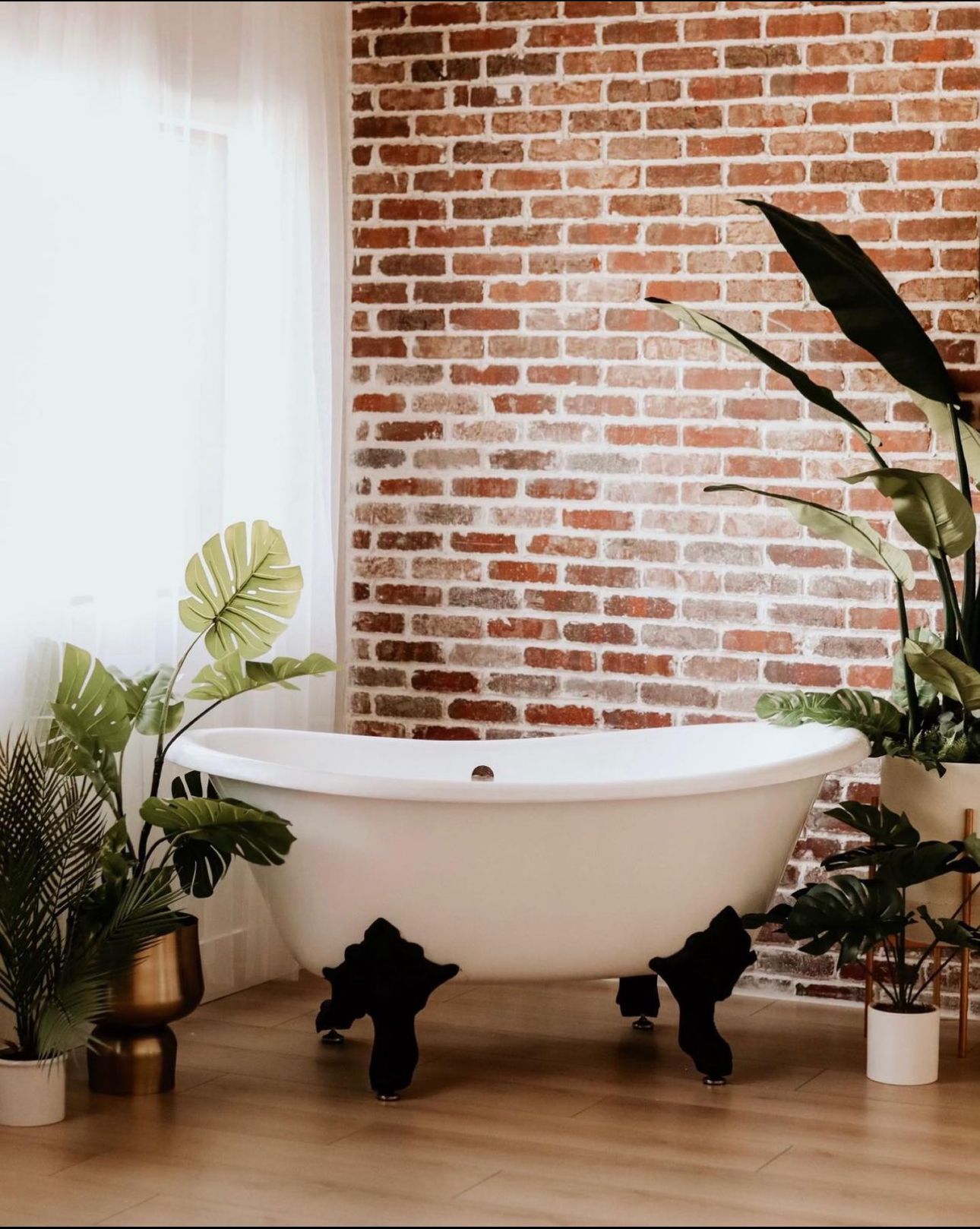 Classic clawfoot bathtub with tropical plants against exposed brick wall in modern bathroom.