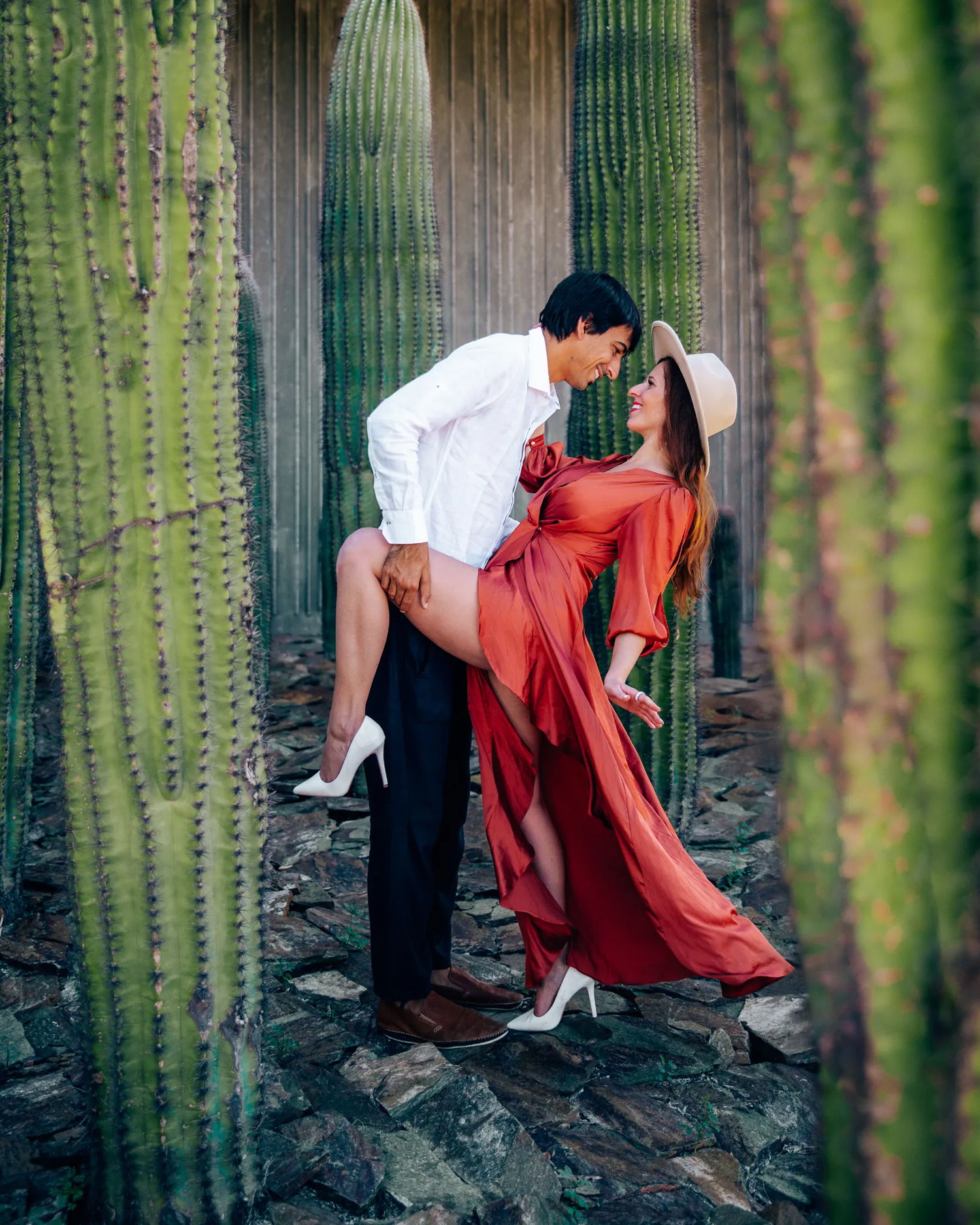 Stylish couple in romantic pose among tall desert cactus in dramatic lighting.