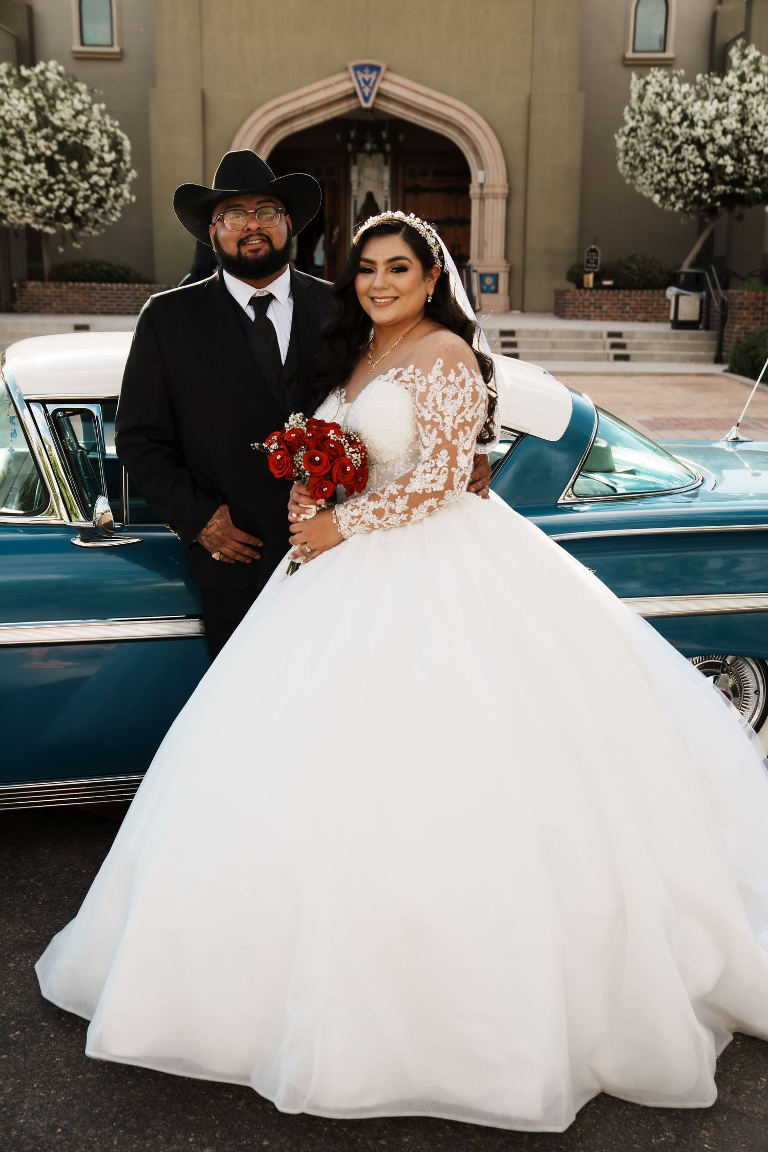 Bride in white ball gown with lace sleeves holds red bouquet beside groom in black cowboy hat near vintage teal car.
