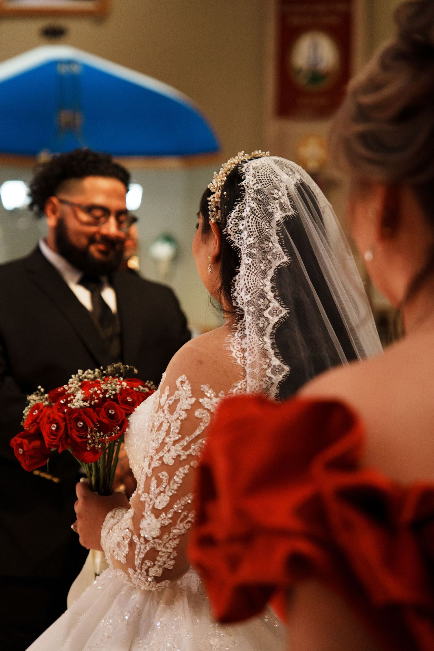 Bride in lace wedding gown holding red rose bouquet faces groom in black suit during church ceremony.