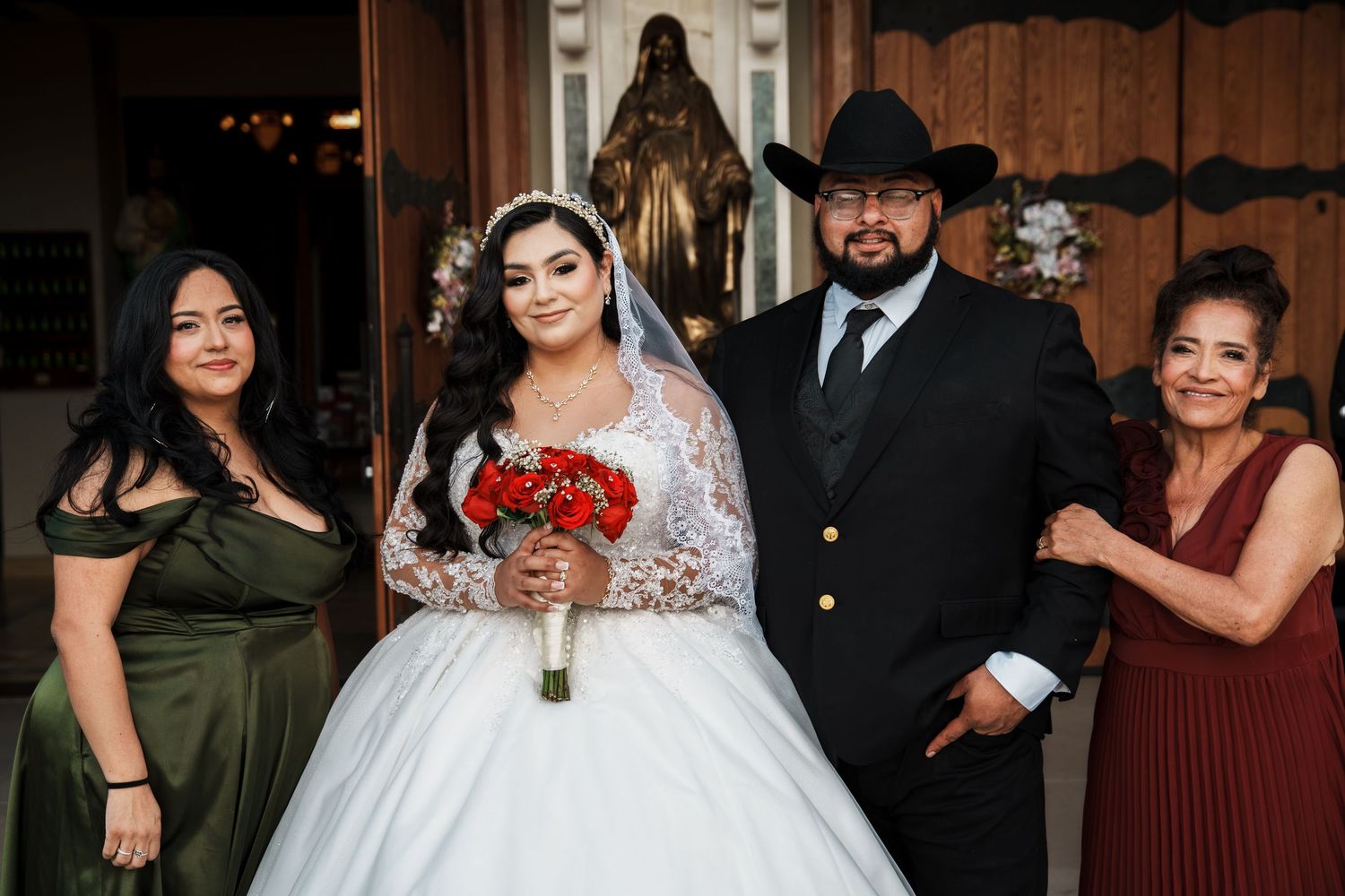 Bride in white ball gown holding red bouquet poses with groom in cowboy hat and two women in church wedding photo.