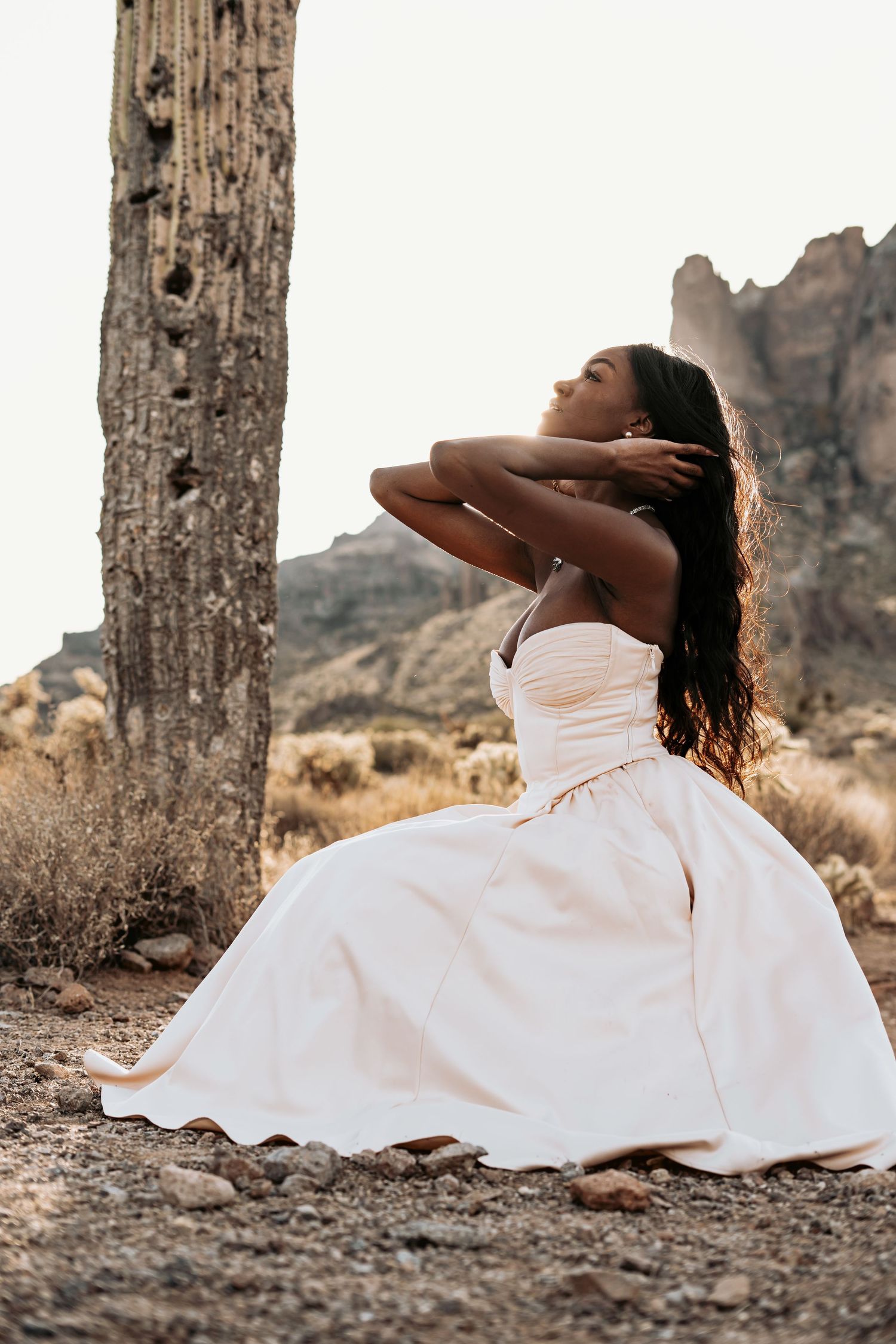 A woman in a flowing white dress poses gracefully against a rugged desert landscape with mountains in the background.