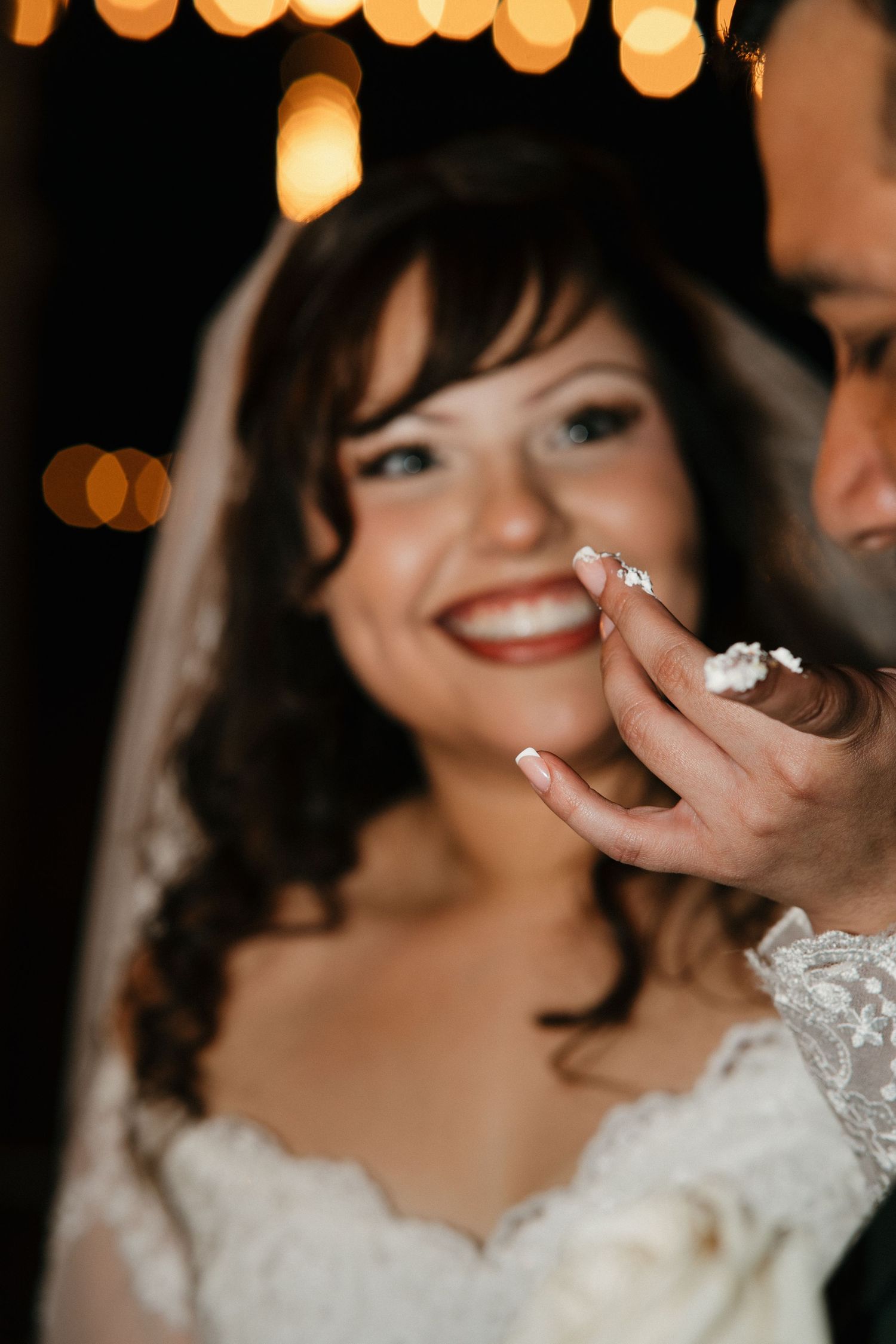 Smiling bride in lace wedding dress and veil with groom feeding her wedding cake at romantic reception.