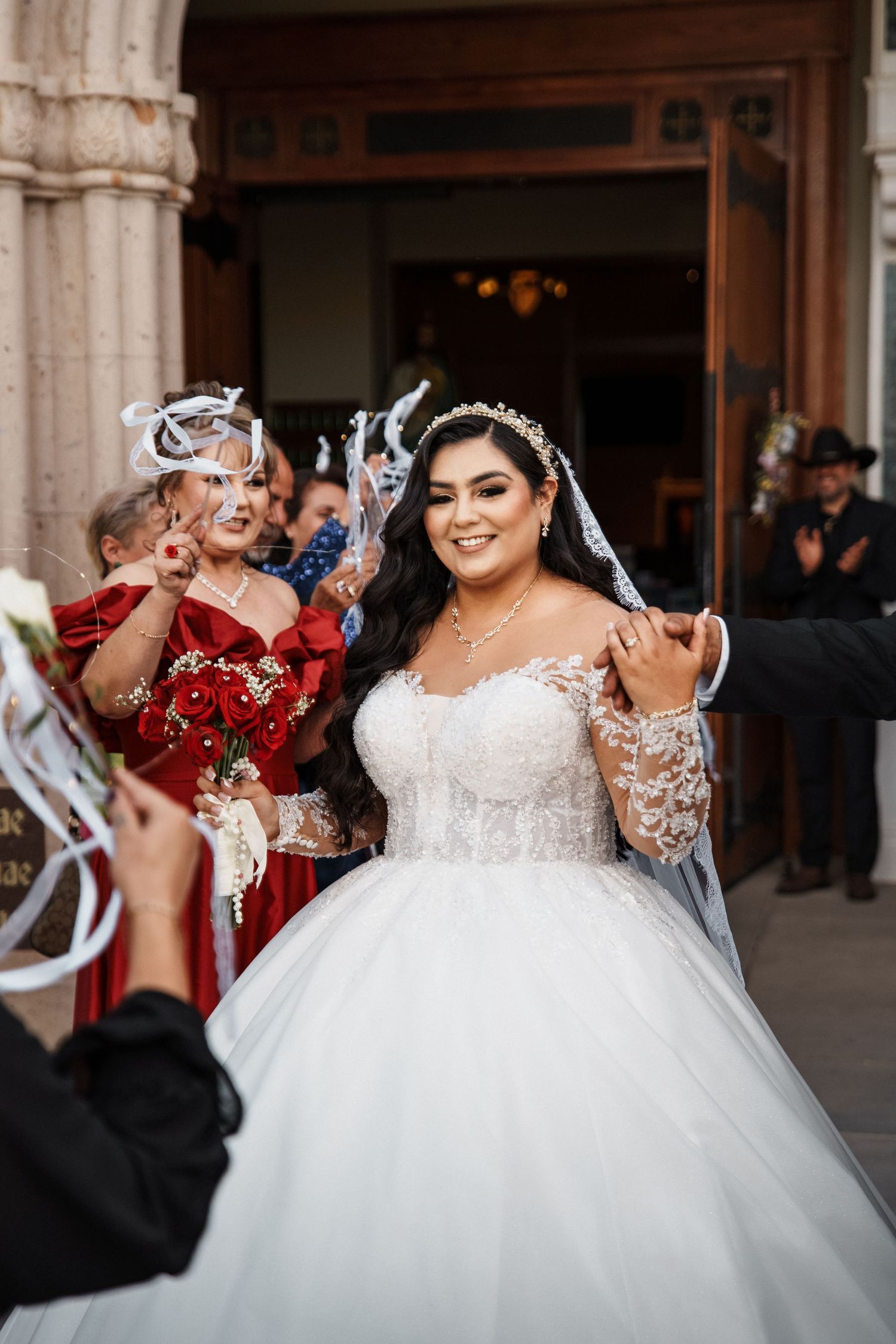 Bride in white lace ball gown smiles joyfully outside church surrounded by guests in red dresses holding ribbon wands.