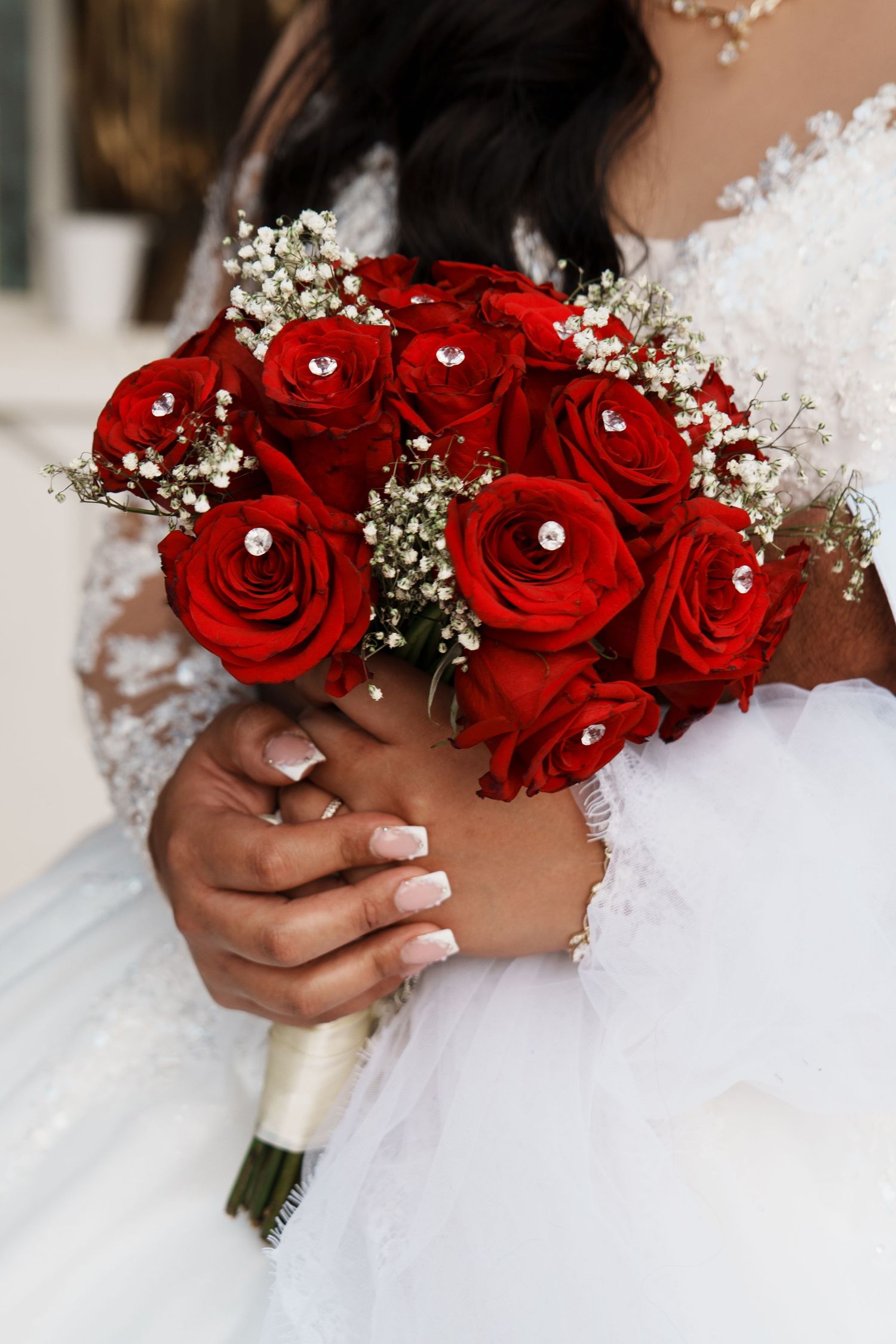 Bride holding a bouquet of red roses with rhinestones and baby's breath in a white lace wedding dress.