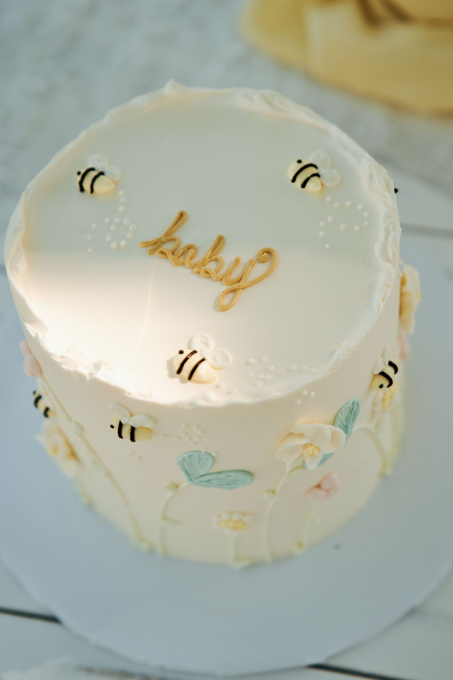 White baby shower cake with gold 'baby' script, bumble bee decorations, and floral accents on frosted tiered design.