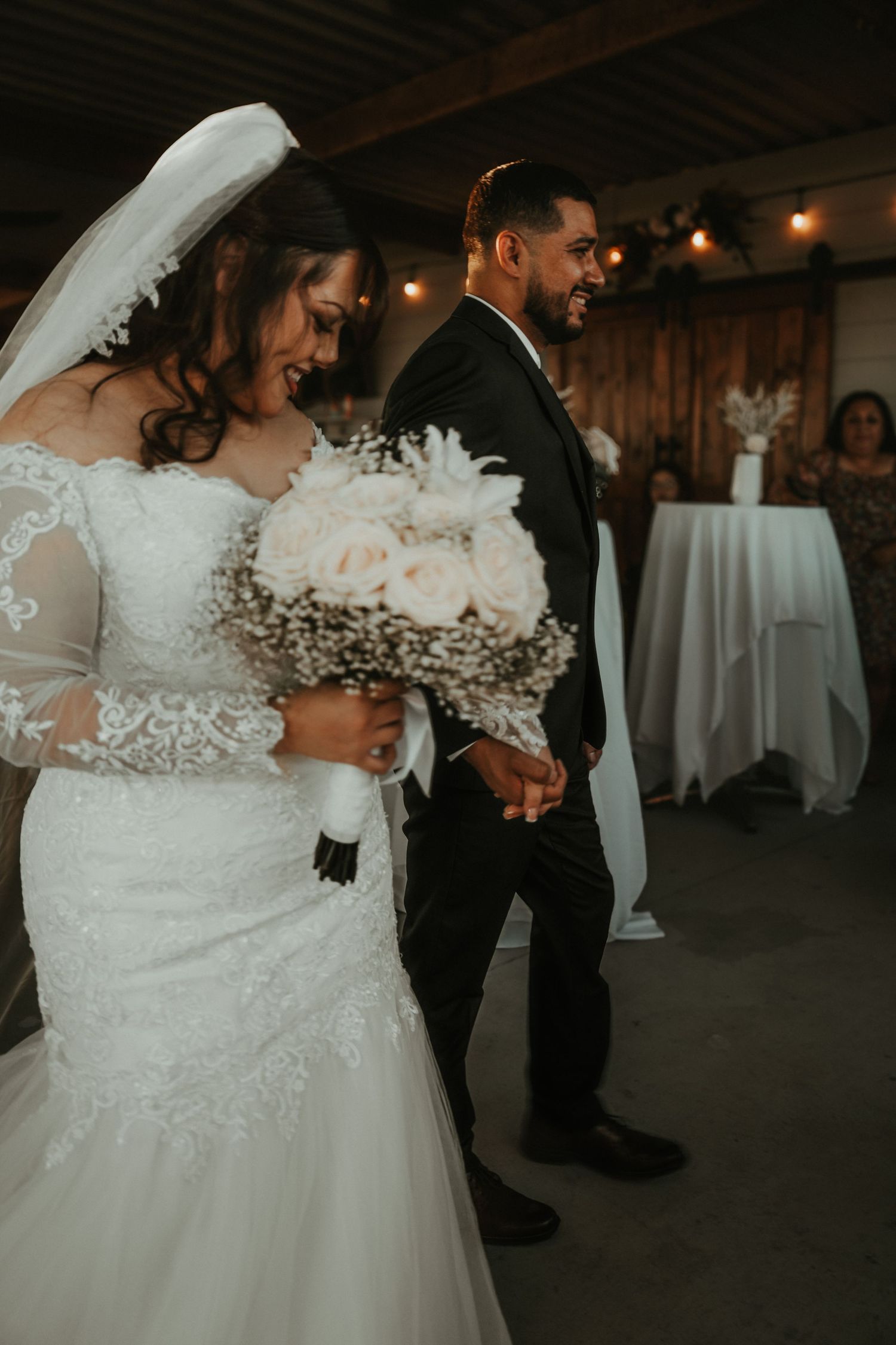 Bride in lace wedding gown holding white rose bouquet walks with groom in black suit during indoor wedding ceremony.