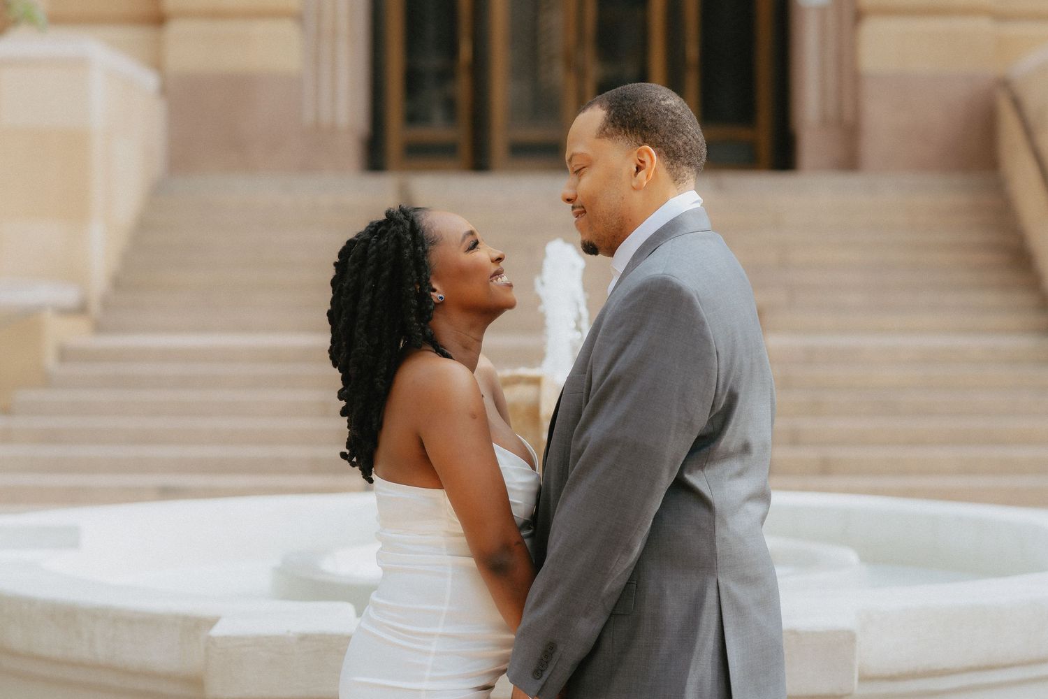 A couple in wedding attire shares a romantic moment by a fountain outside a grand building.