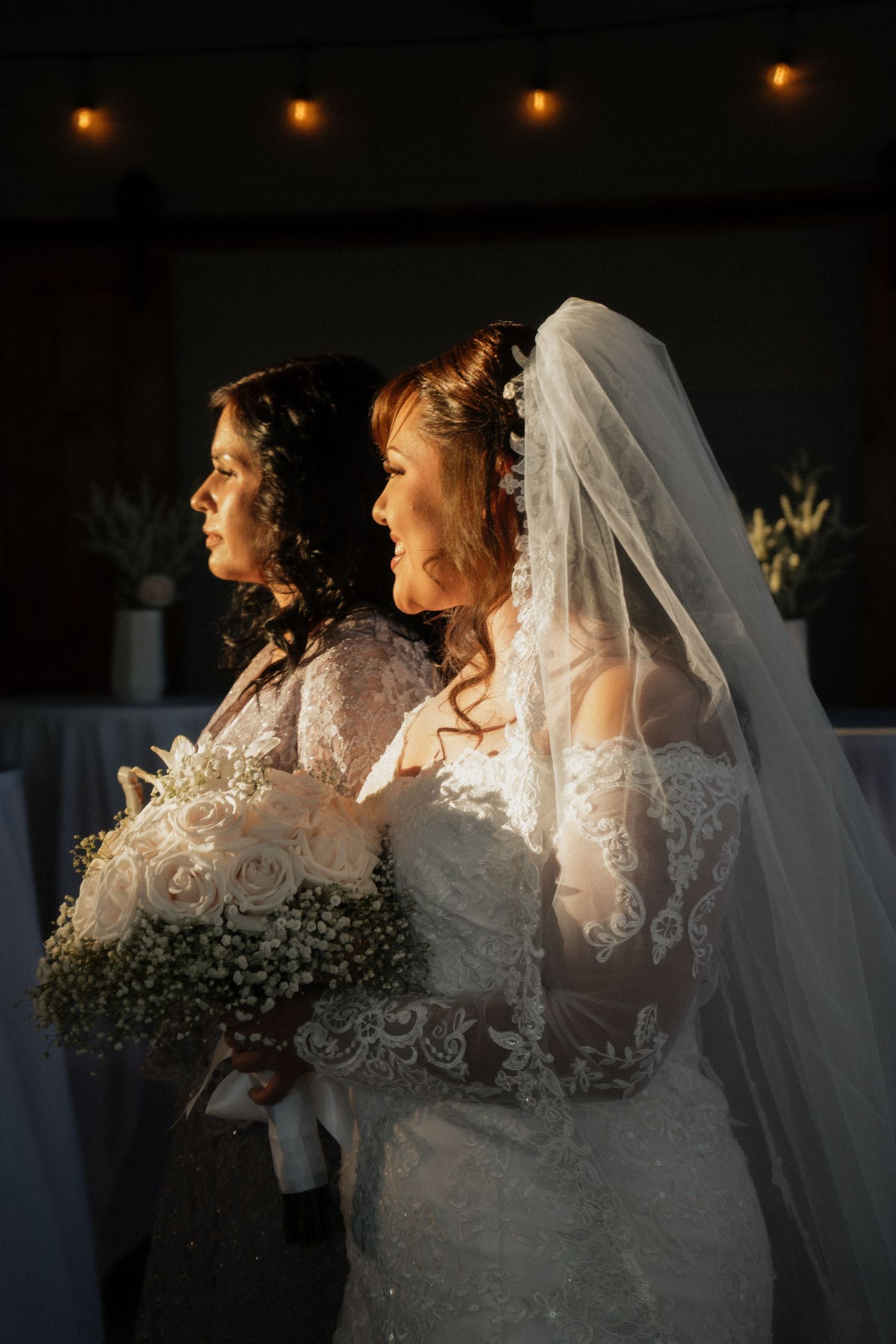 Two brides in white lace gowns stand back-to-back, one holding a bouquet of white roses and baby's breath.