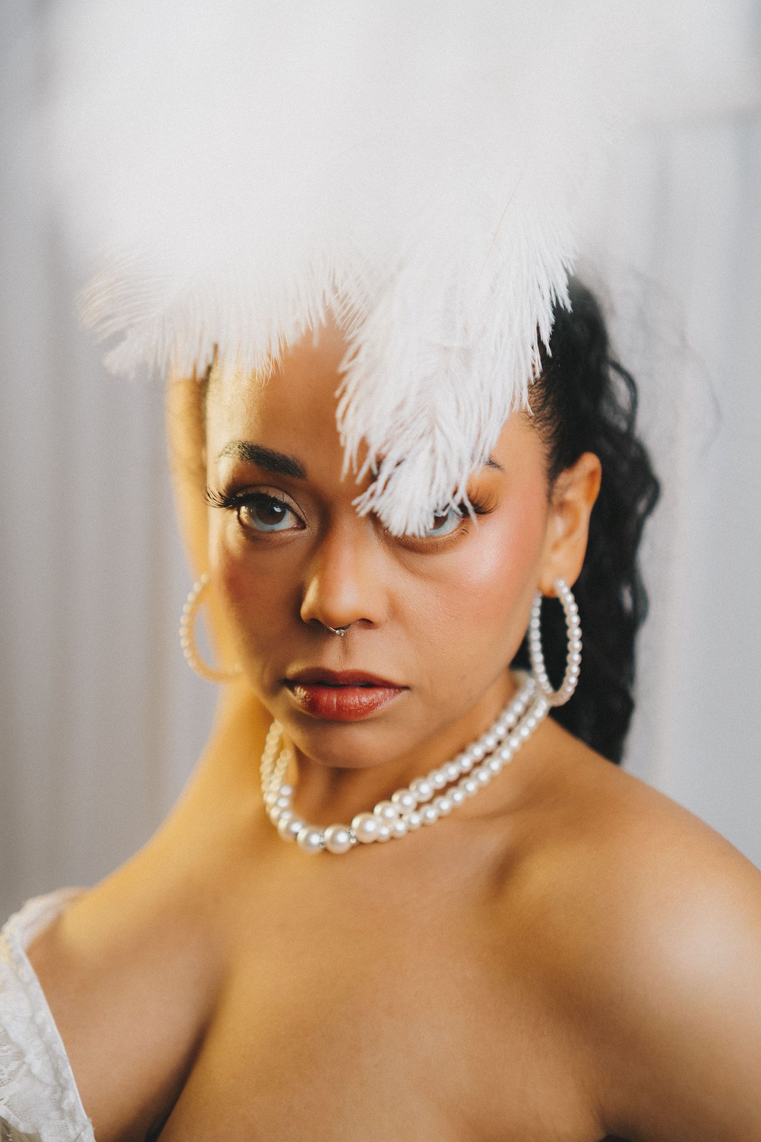 Woman wearing white feather headpiece, pearl necklace, and hoop earrings poses confidently against light background.