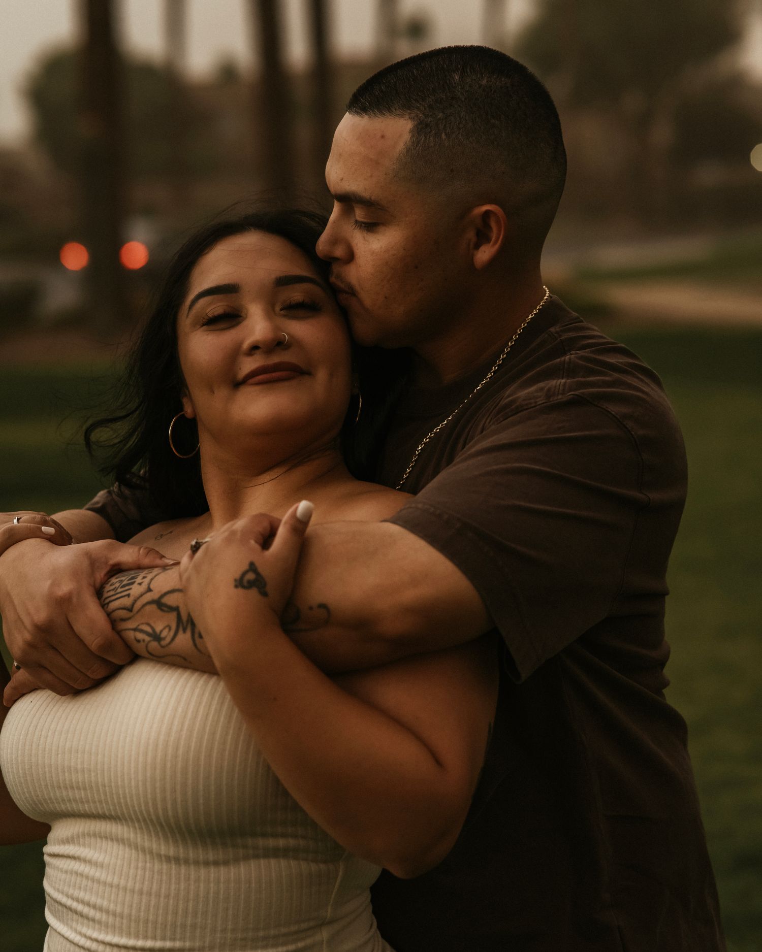 A couple embraces tenderly at dusk, the man kissing the woman's cheek as she smiles warmly in a park setting.