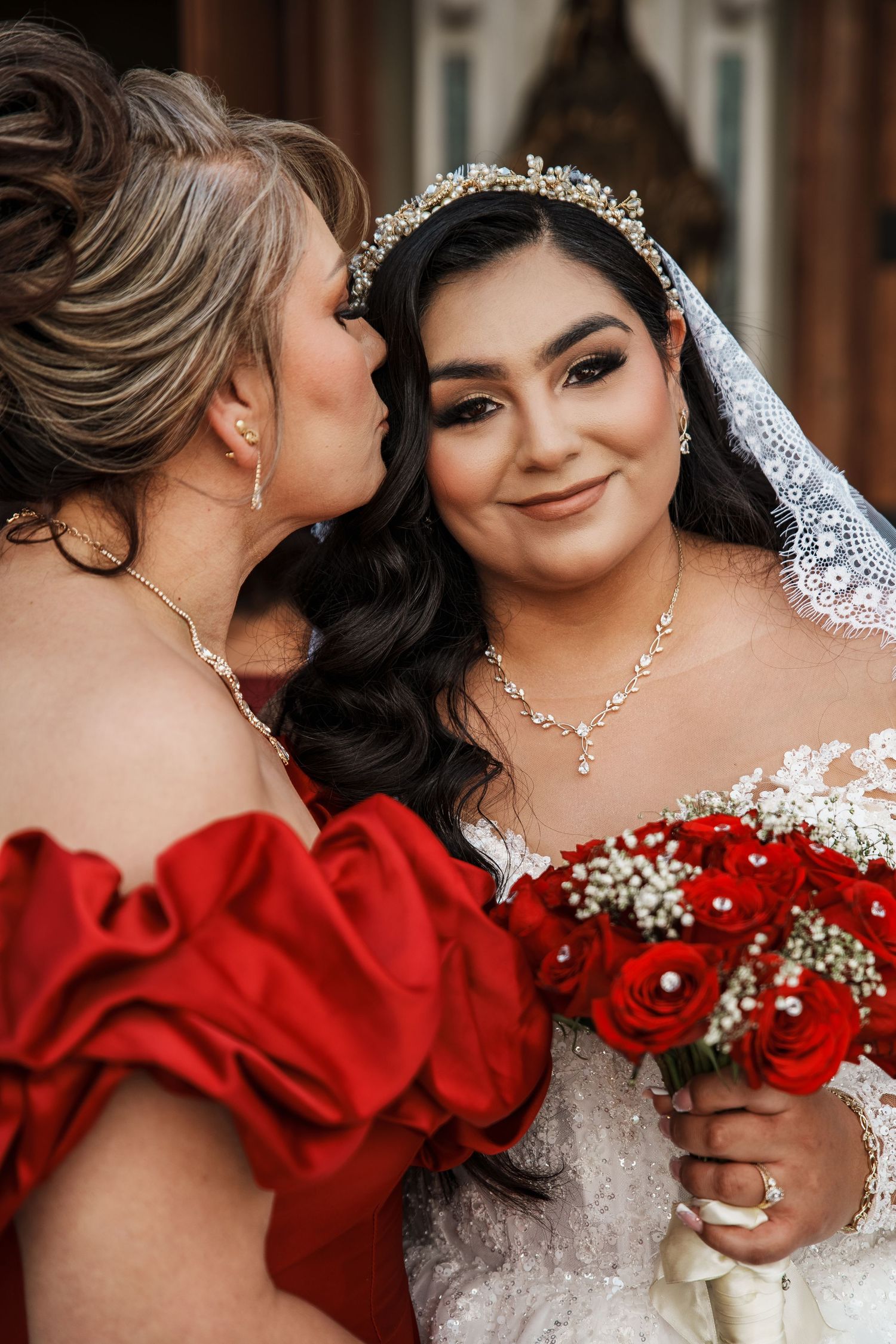 A bride in white lace gown holds red roses as a woman in red dress kisses her cheek at a wedding.