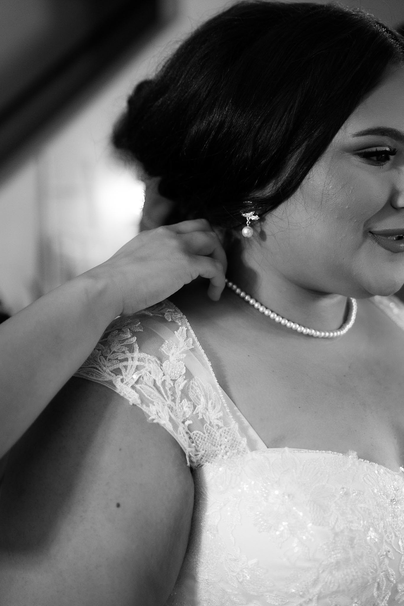 Black and white photo of a bride in lace gown and pearl necklace having her earring adjusted on her wedding day.