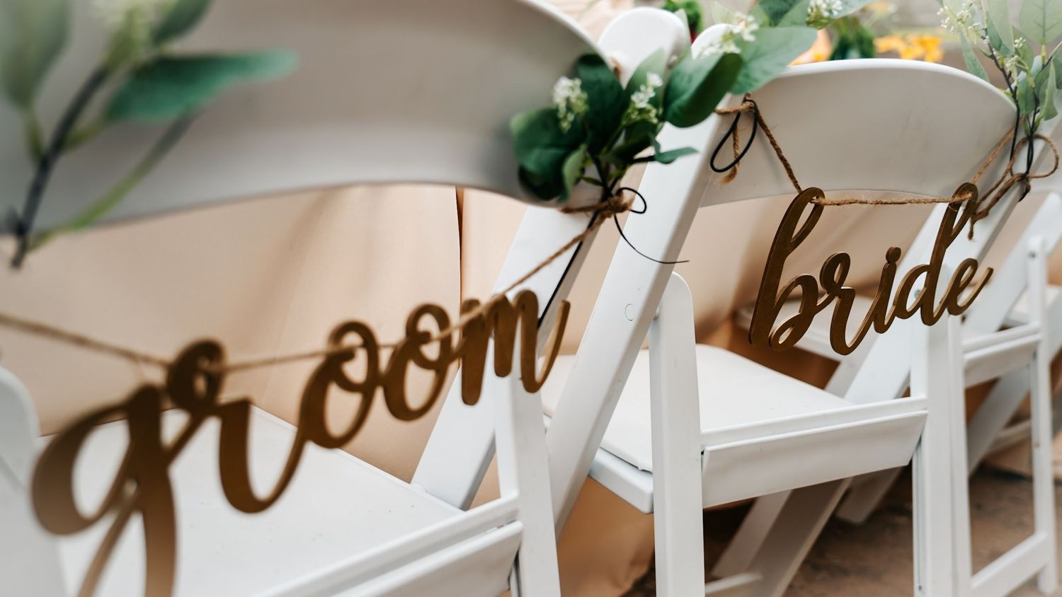 Elegant wooden groom and bride chair signs hanging on white folding chairs at a wedding ceremony.