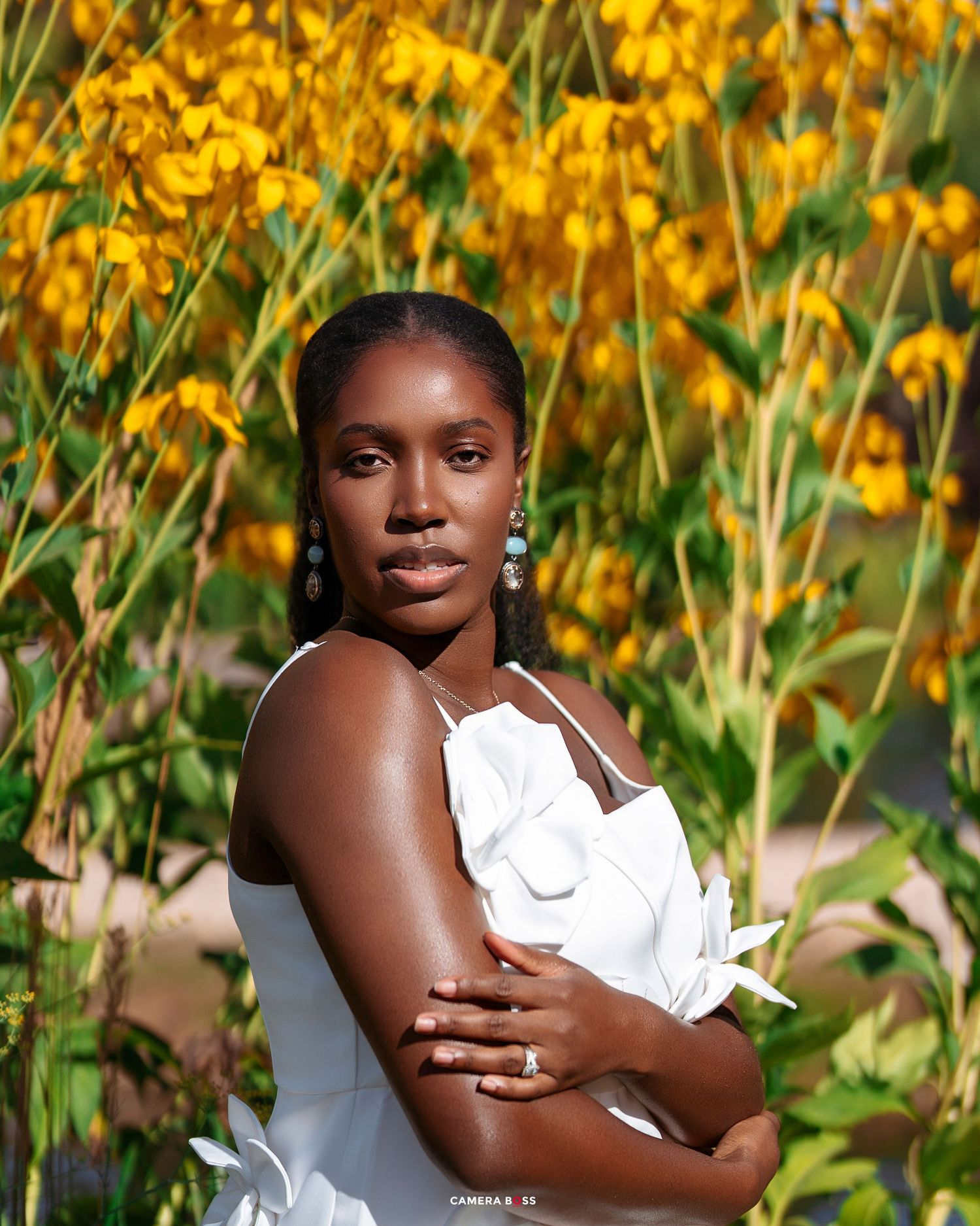 A young Black woman in a white dress poses gracefully among vibrant yellow flowers in a sunny garden.