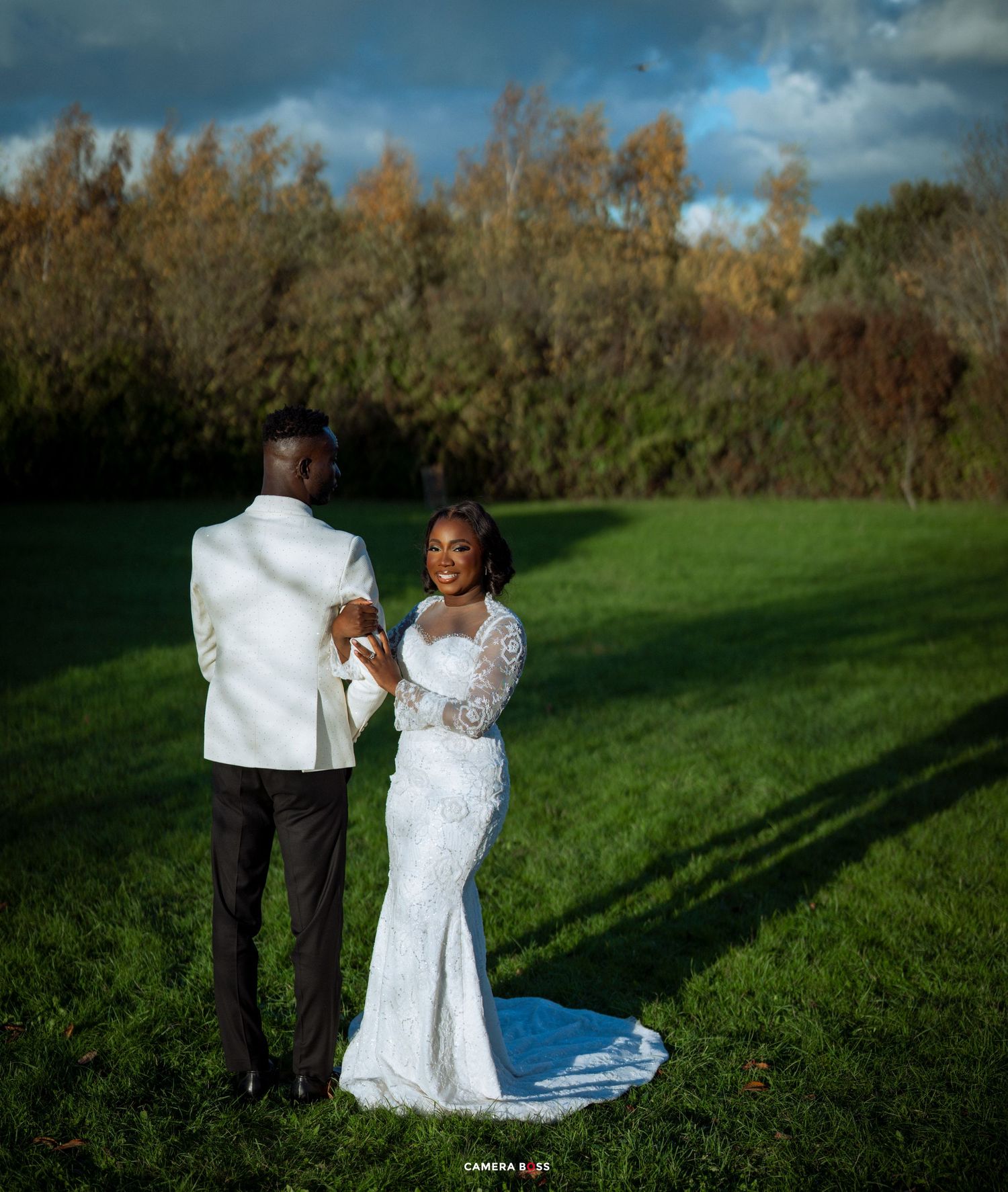 Newlyweds dance together in a grassy field on a sunny autumn day with trees in the background.