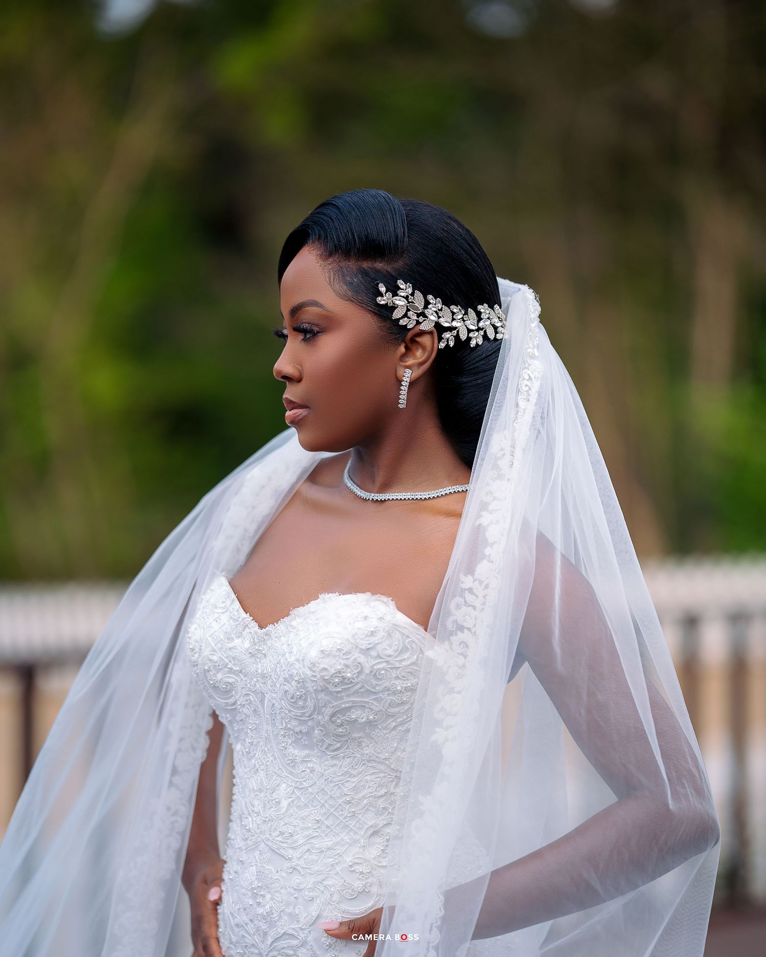 Beautiful bride in strapless lace wedding gown with crystal hair accessory and flowing white veil against garden backdrop.