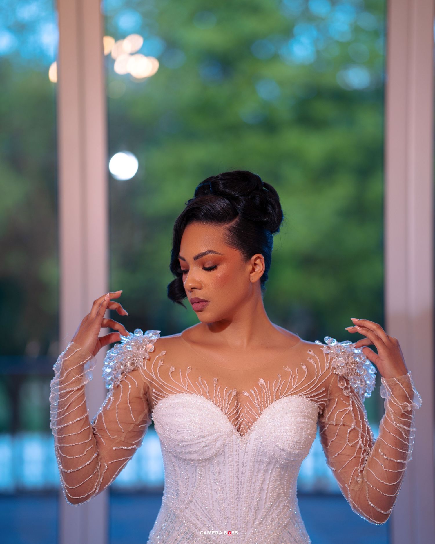 A bride in an elegant white wedding gown with sheer sleeves and feather details poses with closed eyes in romantic lighting.