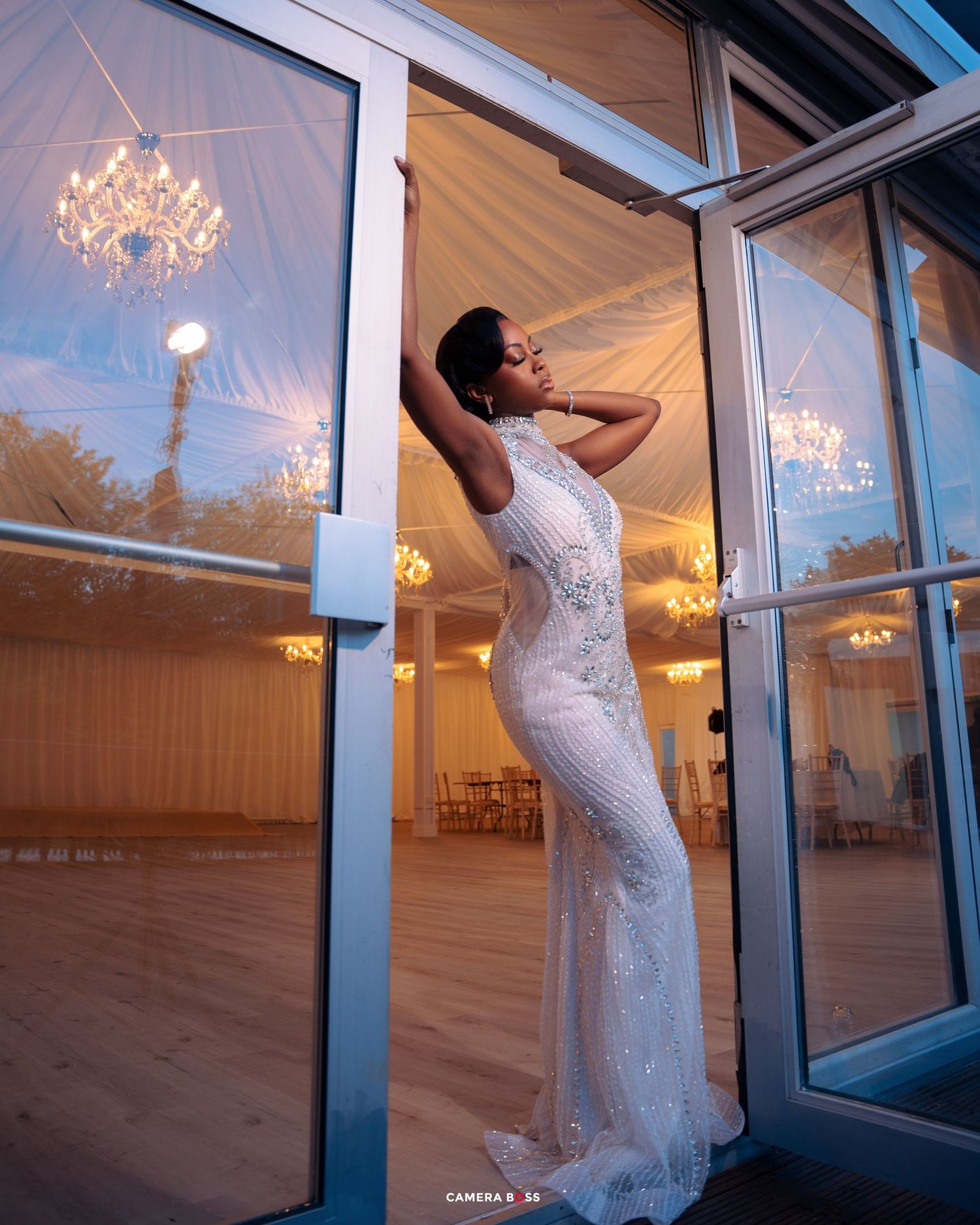 Elegant lace wedding gown silhouetted against glass doors at sunset with crystal chandeliers glowing in the reflection.