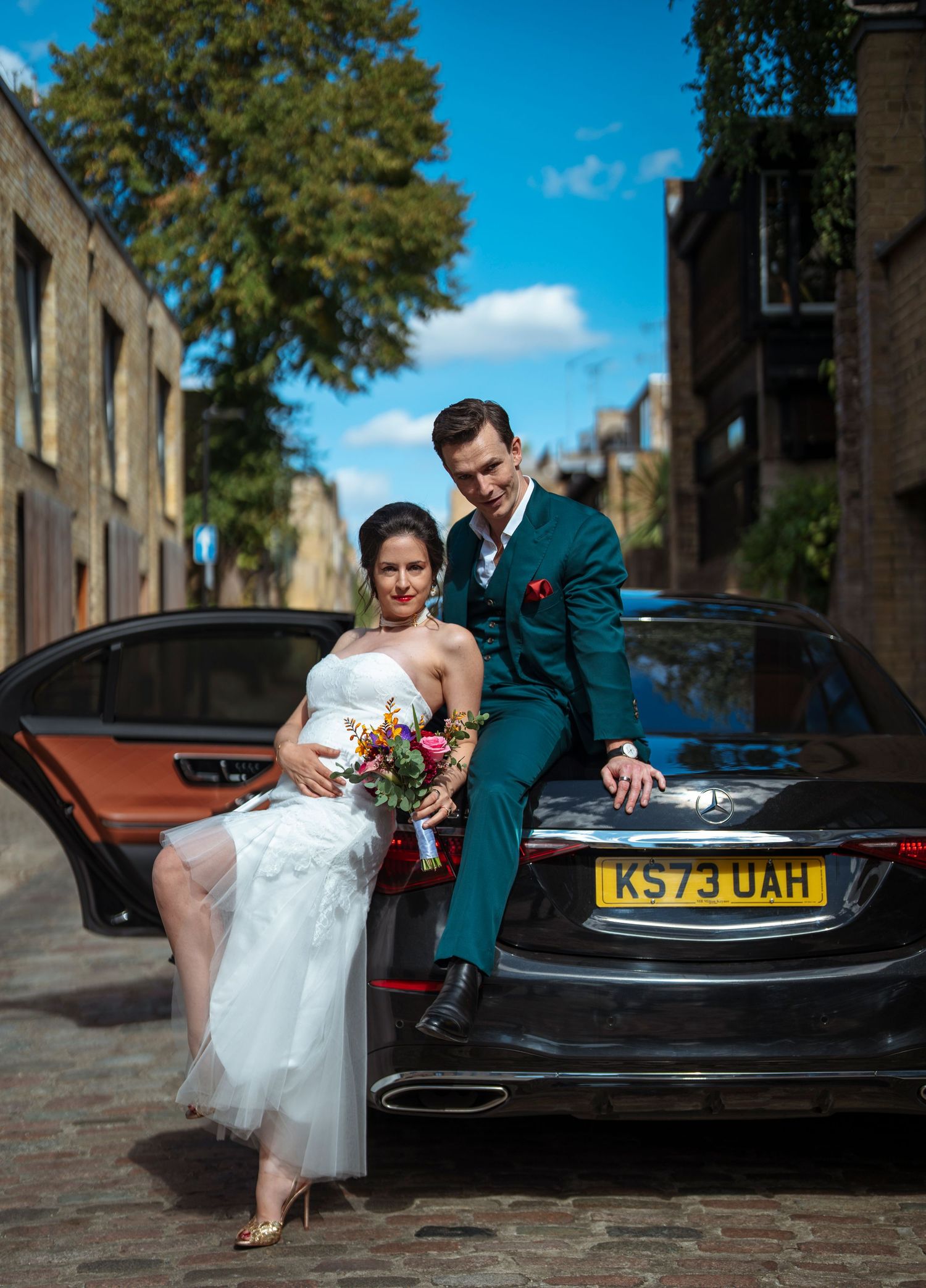 Bride and groom posing elegantly by a black Mercedes on a cobblestone London street on a sunny wedding day.