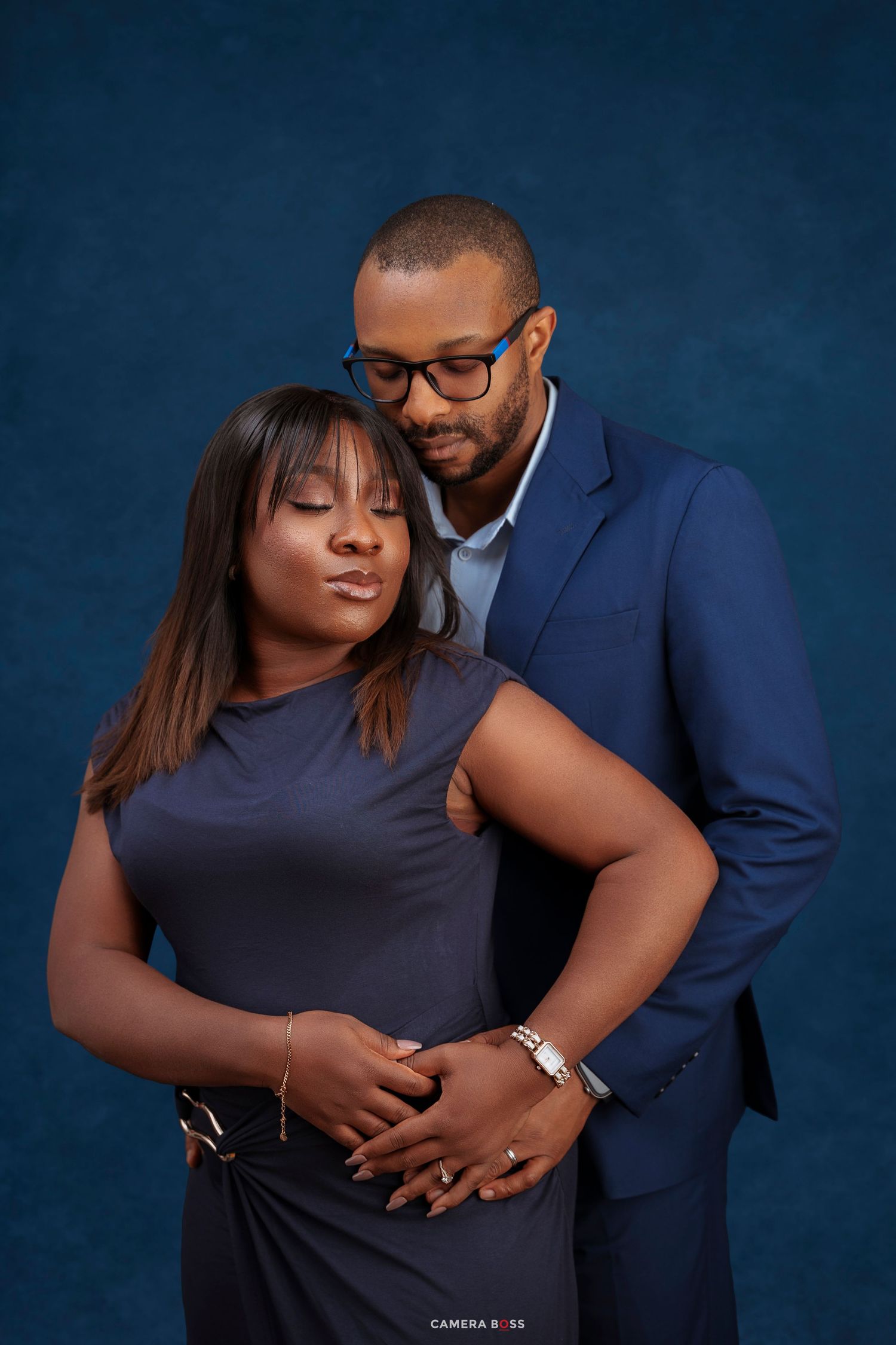 A couple in coordinating navy outfits share an intimate embrace against a deep blue studio background.