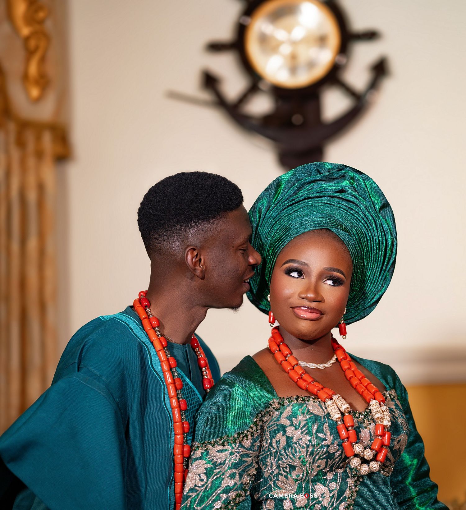 A couple in traditional Nigerian attire with teal clothing and coral beaded jewelry share a tender moment together.
