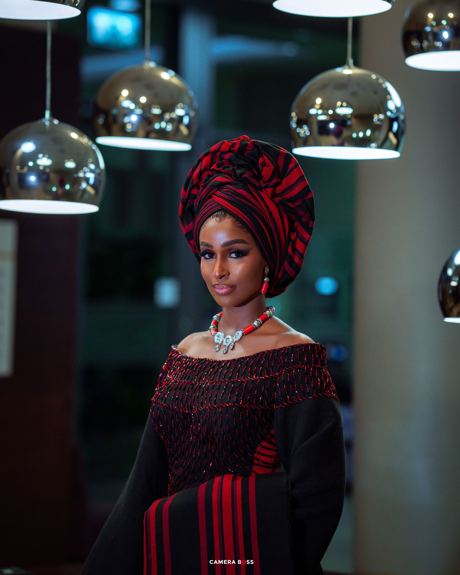 Elegant fashion portrait with a burgundy headwrap and off-shoulder black dress under modern chrome pendant lights.