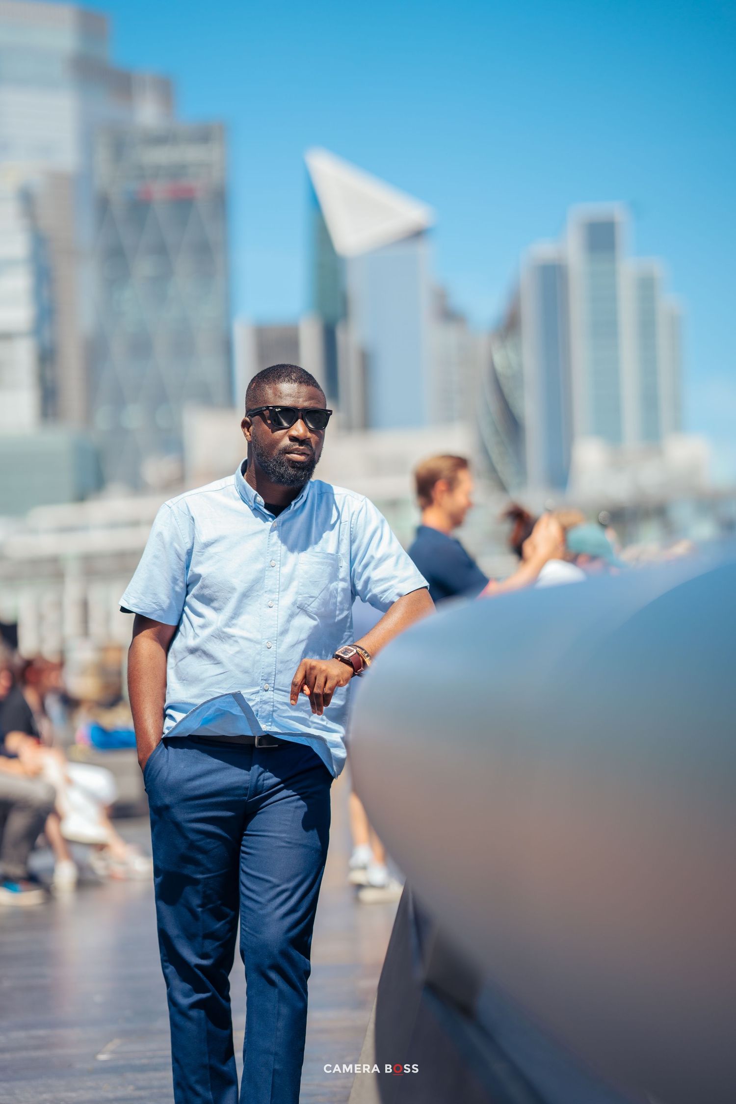 Person in light blue shirt and navy pants stands on urban boardwalk with modern skyscrapers in background.