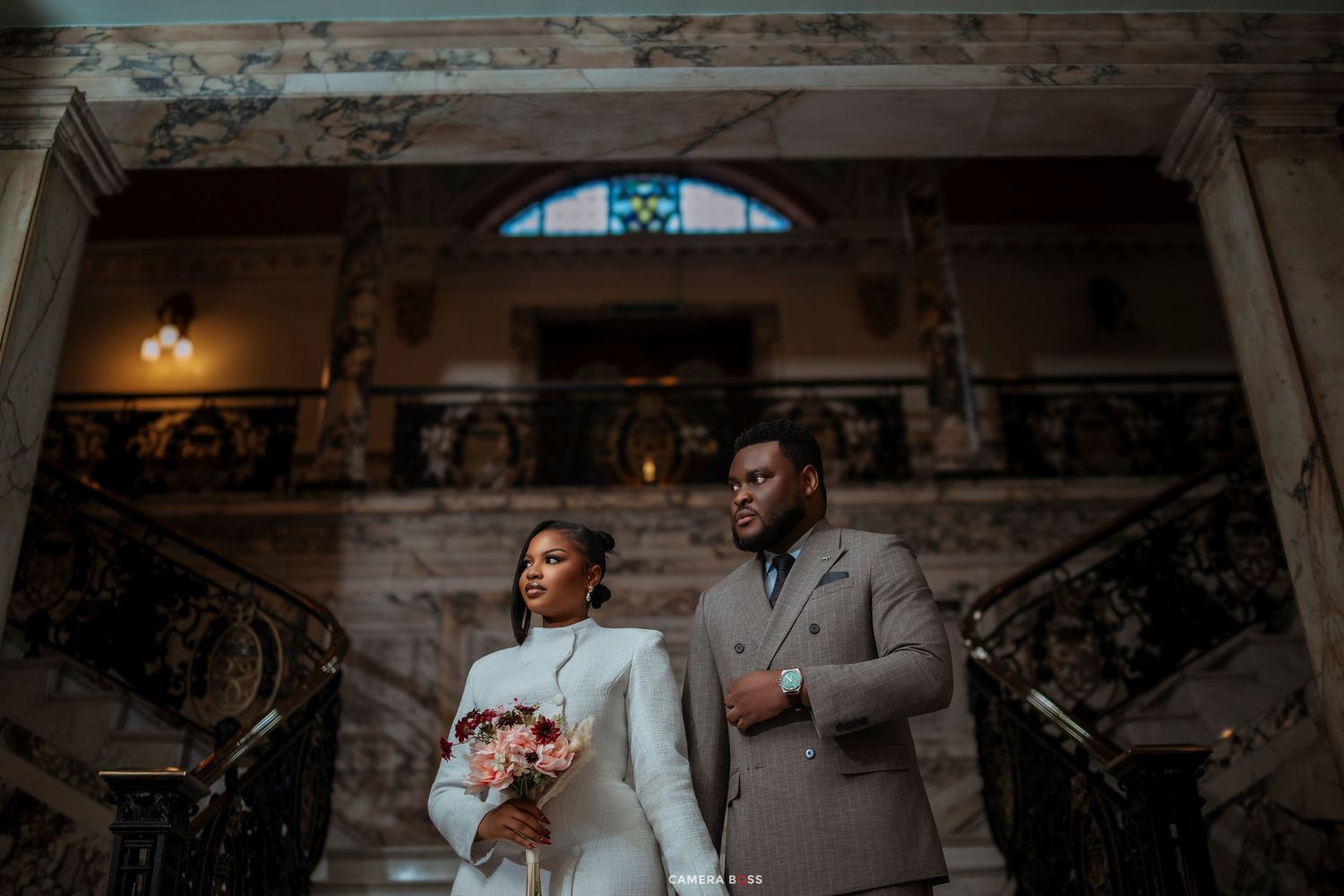Elegant couple in wedding attire poses on ornate staircase, bride holding bouquet in historic venue.