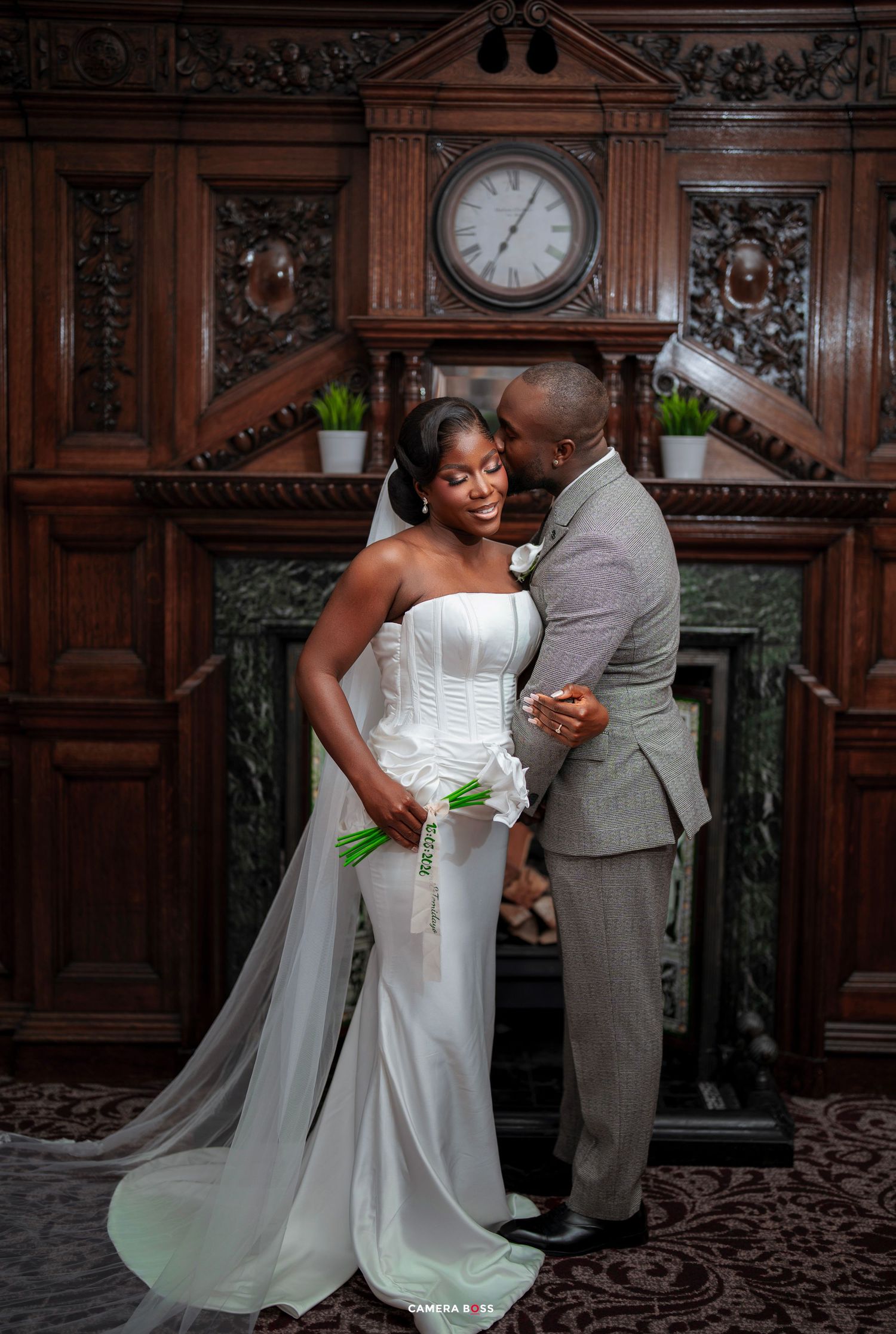 A bride in a white mermaid gown and groom in a gray suit pose romantically before an ornate wooden fireplace.