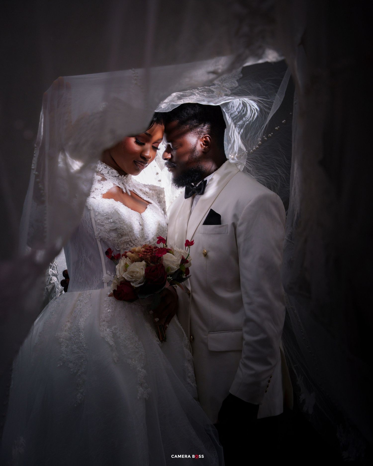 Bride and groom in white wedding attire share romantic moment under flowing veil, holding red and white bouquet.