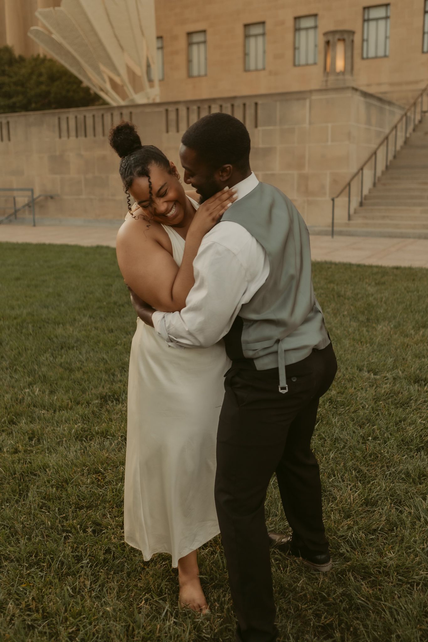 A romantic couple in wedding attire share a tender moment while standing on grass near stone steps.