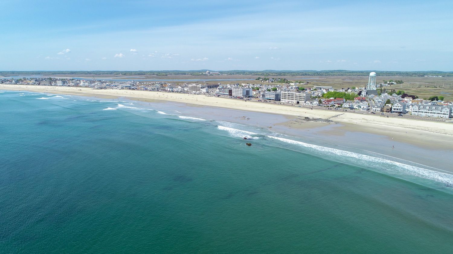 Aerial view of a sandy beach coastline with turquoise ocean waves and a small coastal town under a blue sky.
