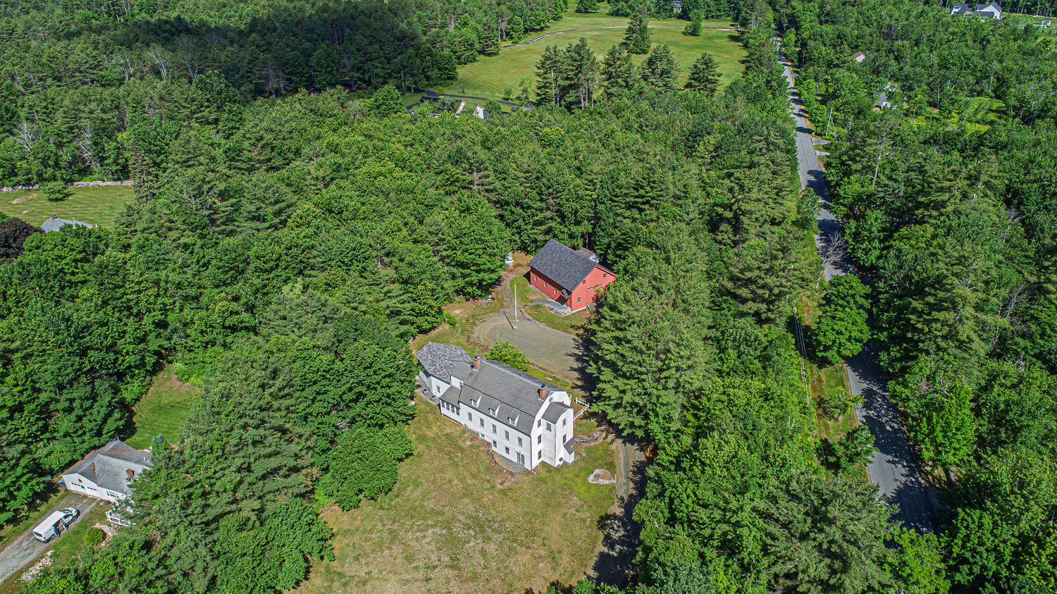 Aerial view of a white colonial house surrounded by dense green trees and forest in a rural New England setting.