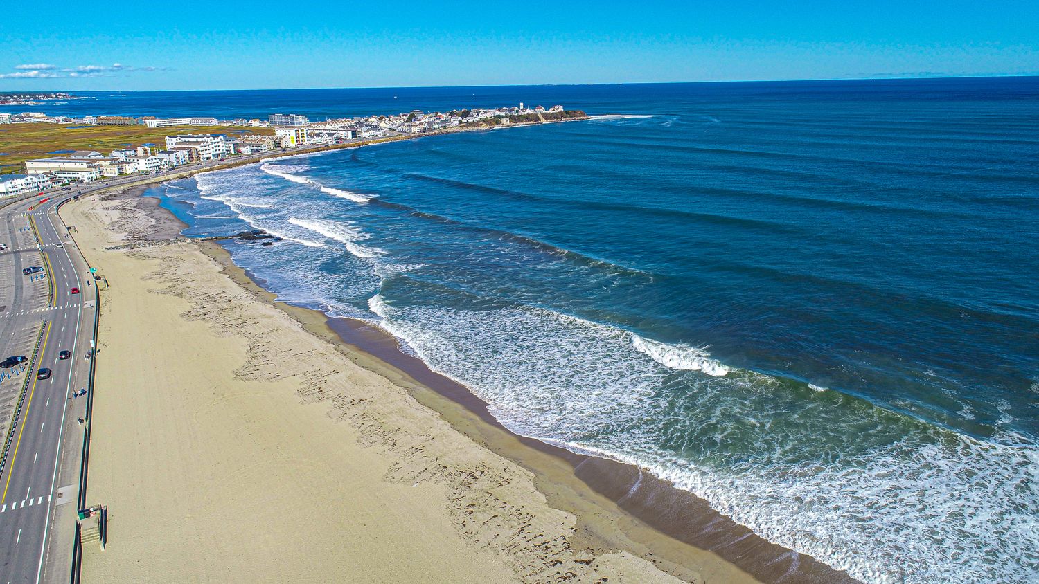 Aerial view of a sandy beach with ocean waves, a coastal road, and a city skyline under a clear blue sky.