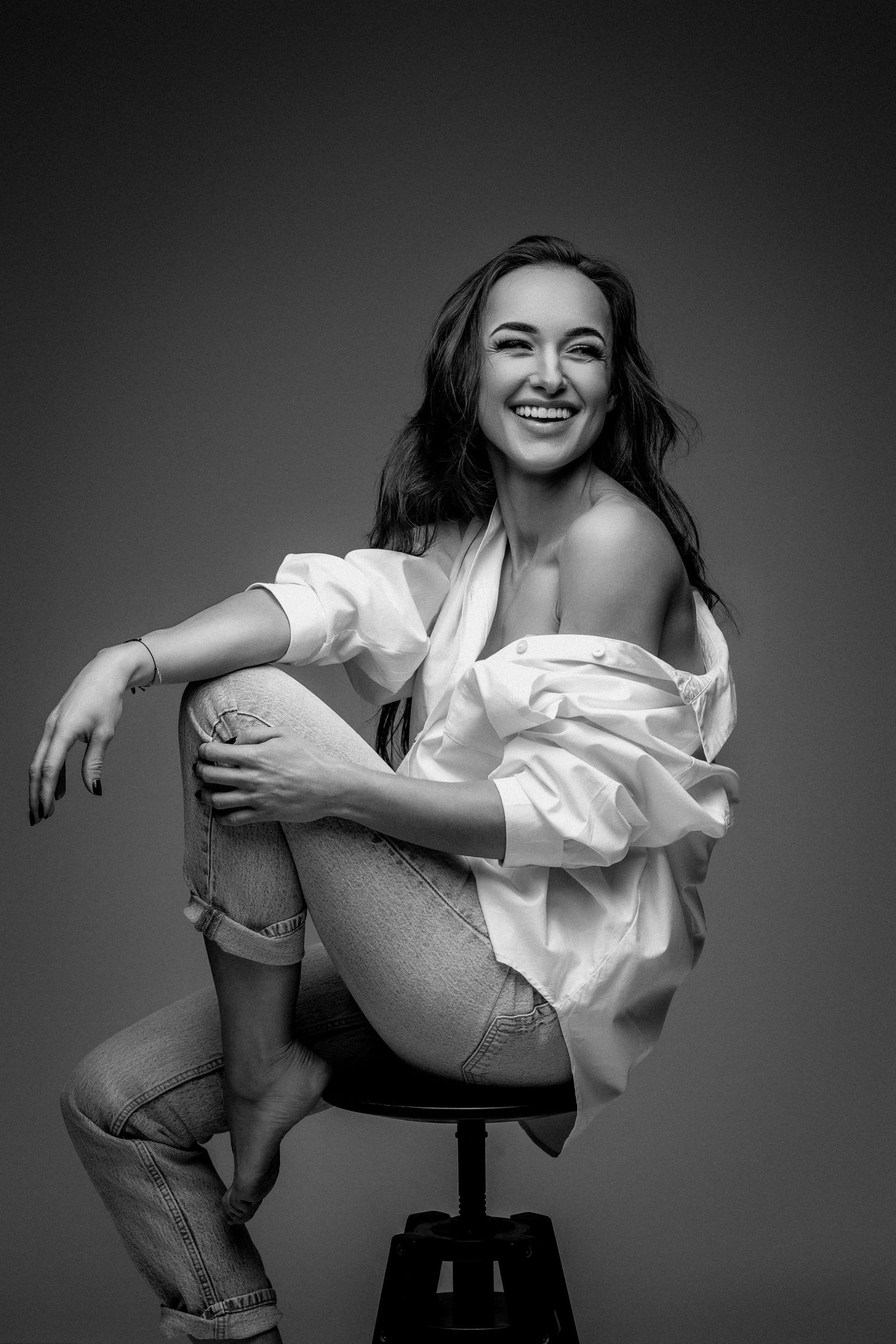 Black and white portrait of model sitting casually on stool in white shirt.