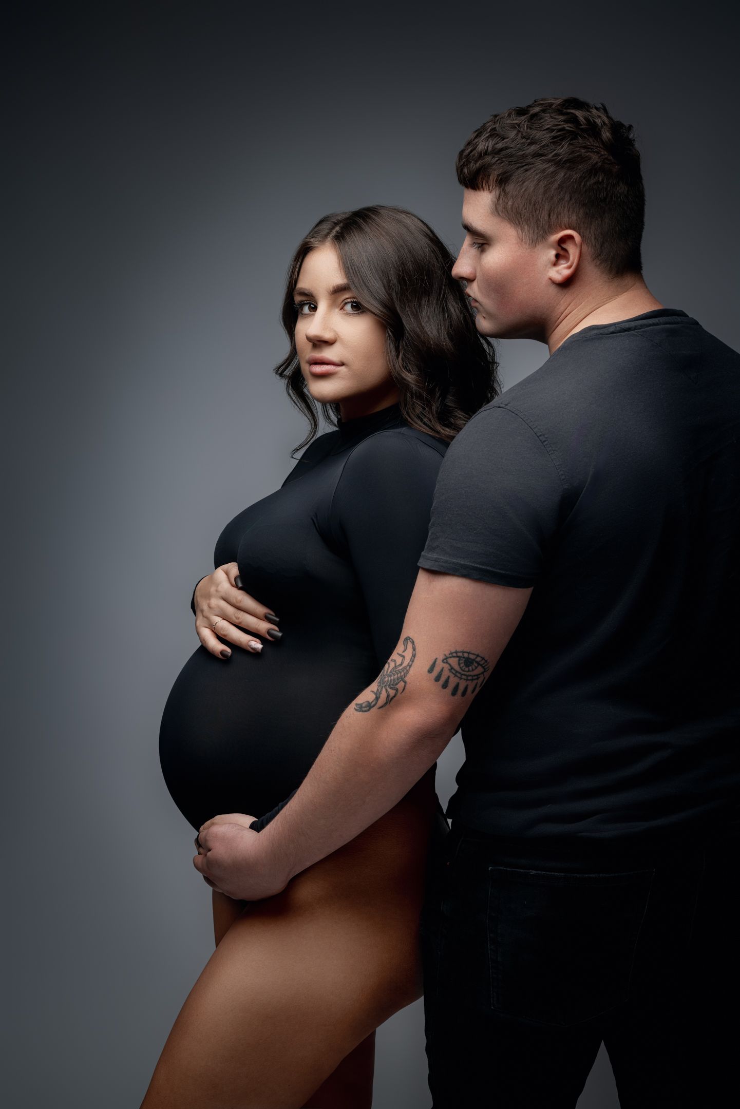 Intimate maternity photo of couple in black clothing against dark background.
