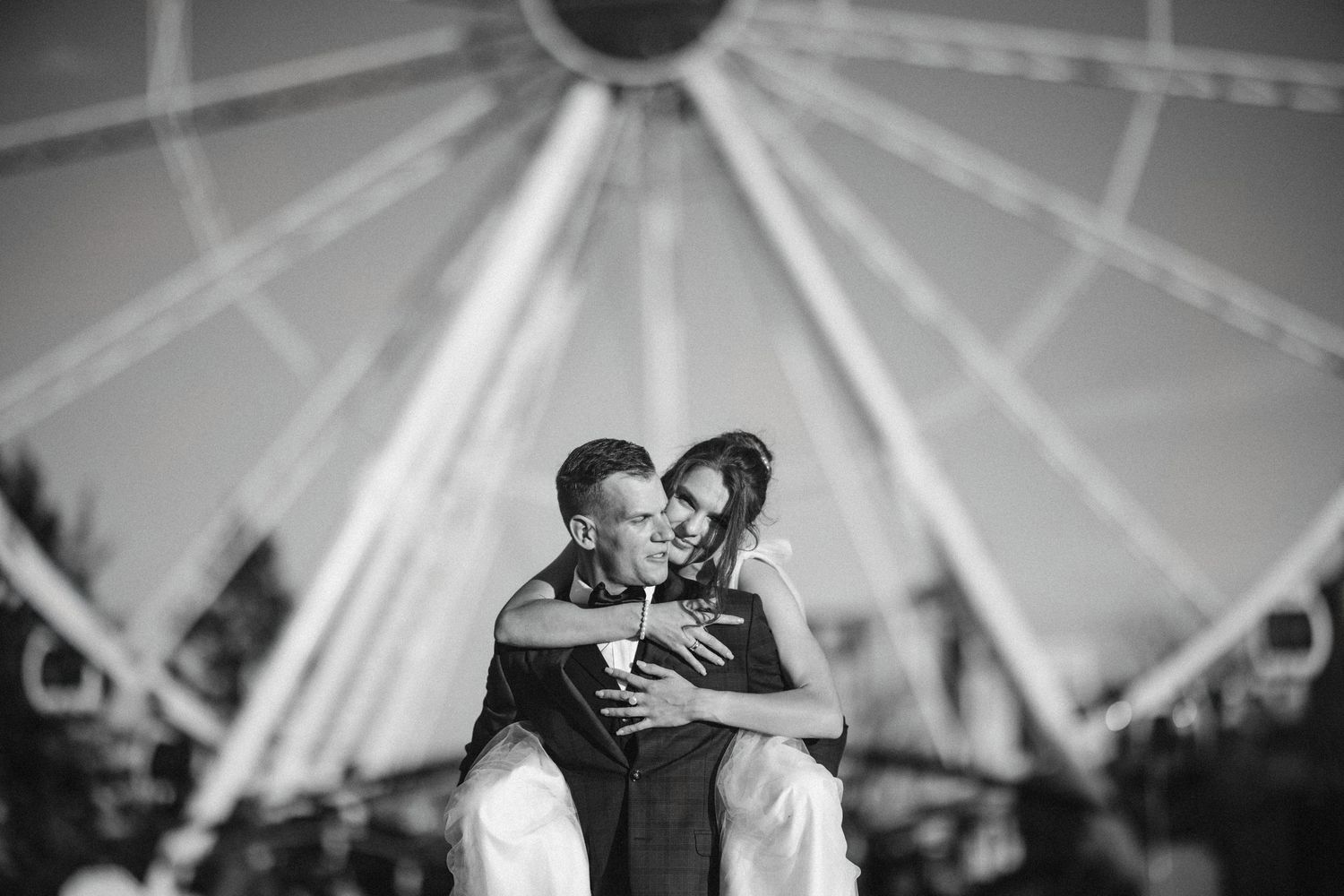 Black and white photo of couple embracing near ferris wheel at night.