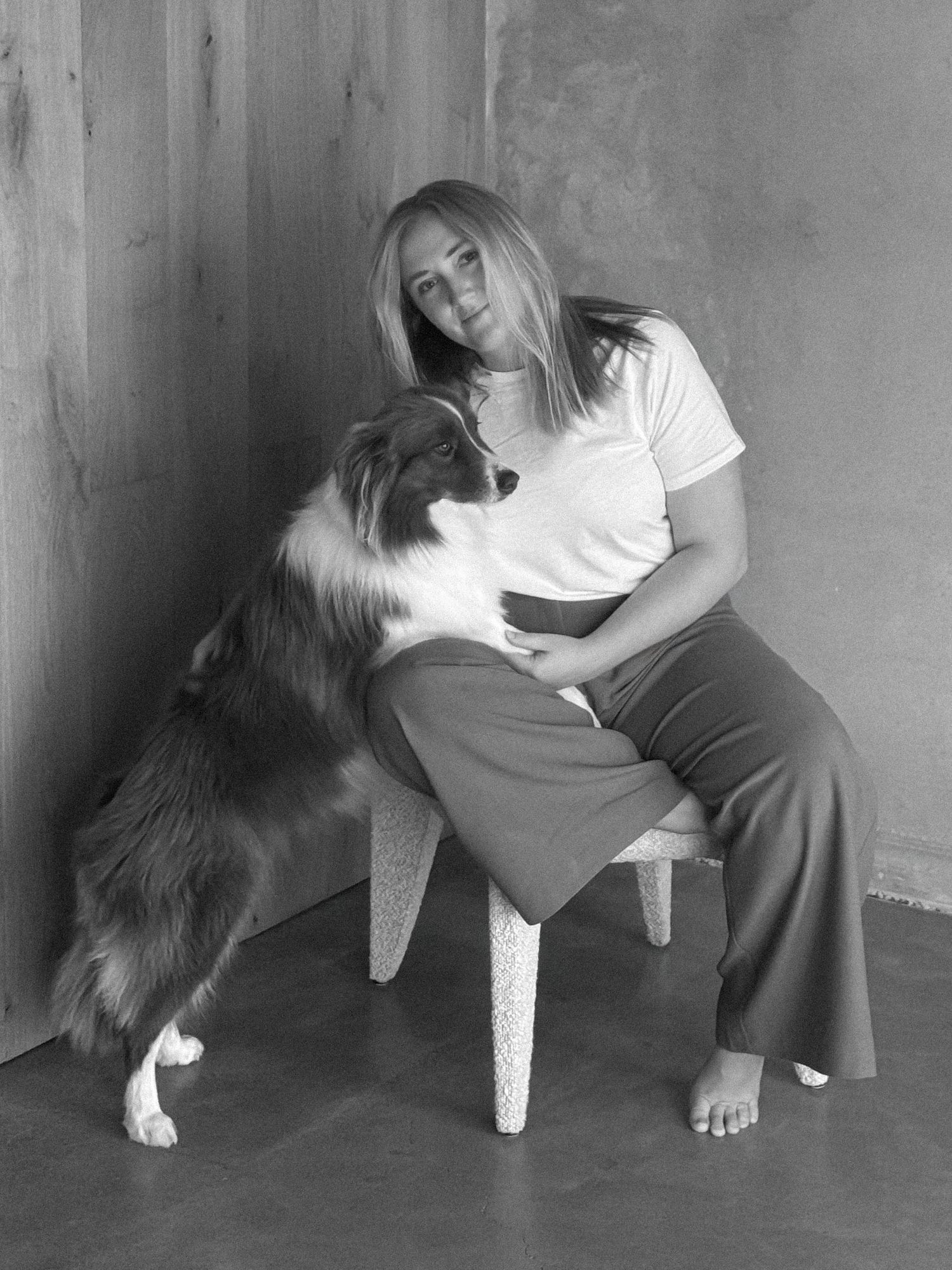 A barefoot woman in a white tee sits on a textured stool with a long-haired dog resting its paws on her lap against a rustic wall.