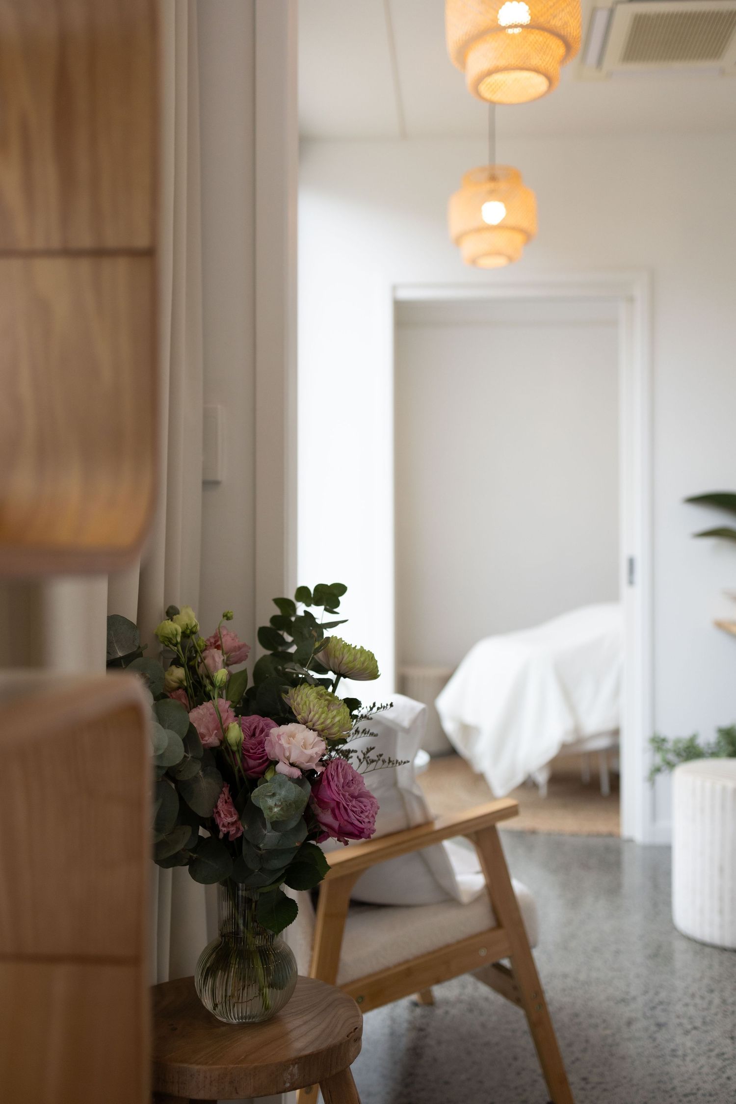 A vase of pink roses sits on a small wooden table in a bright spa reception with woven pendant lights and a treatment room beyond.