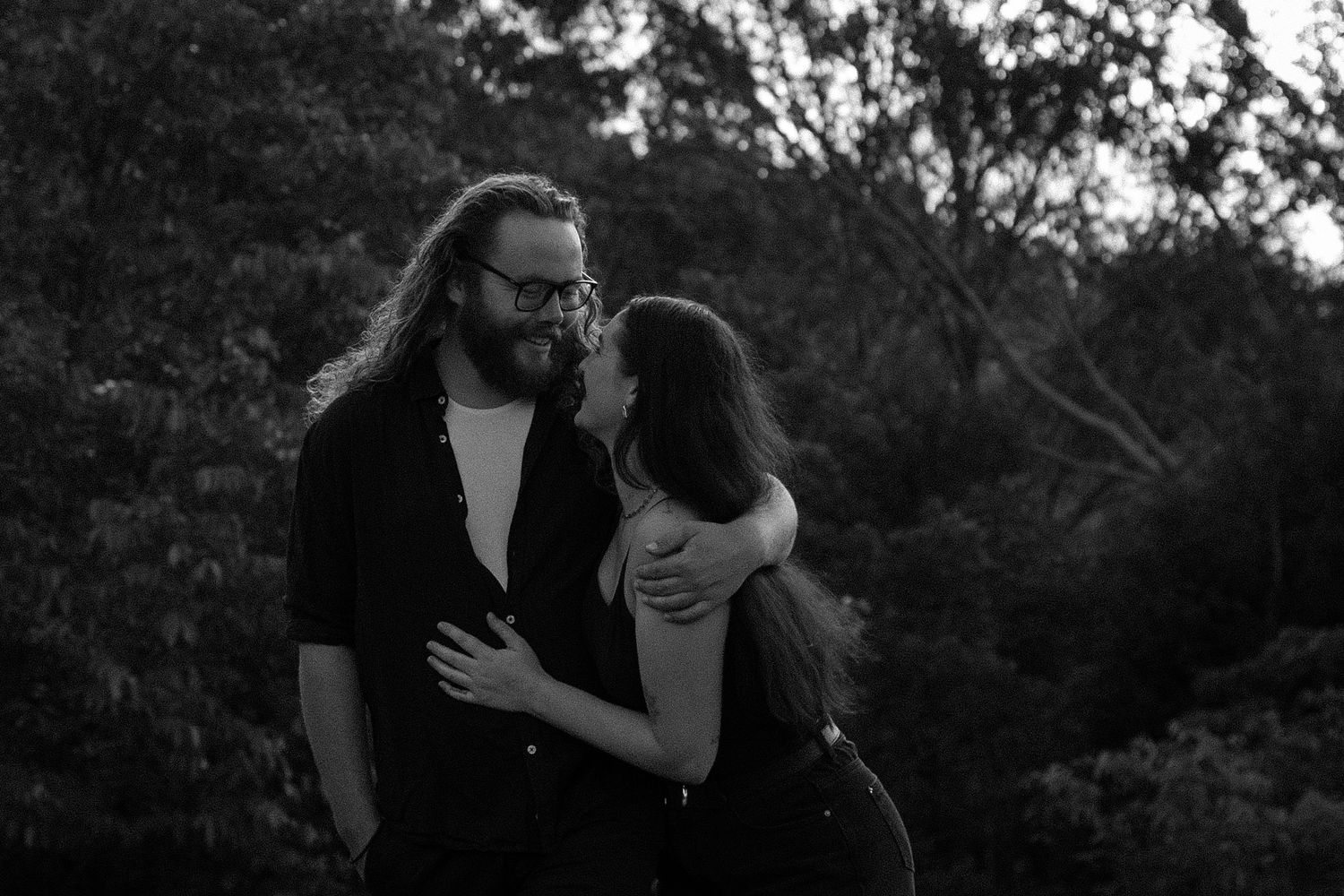 Black and white portrait of a bearded man with long hair and glasses embracing a woman outdoors among dark trees.