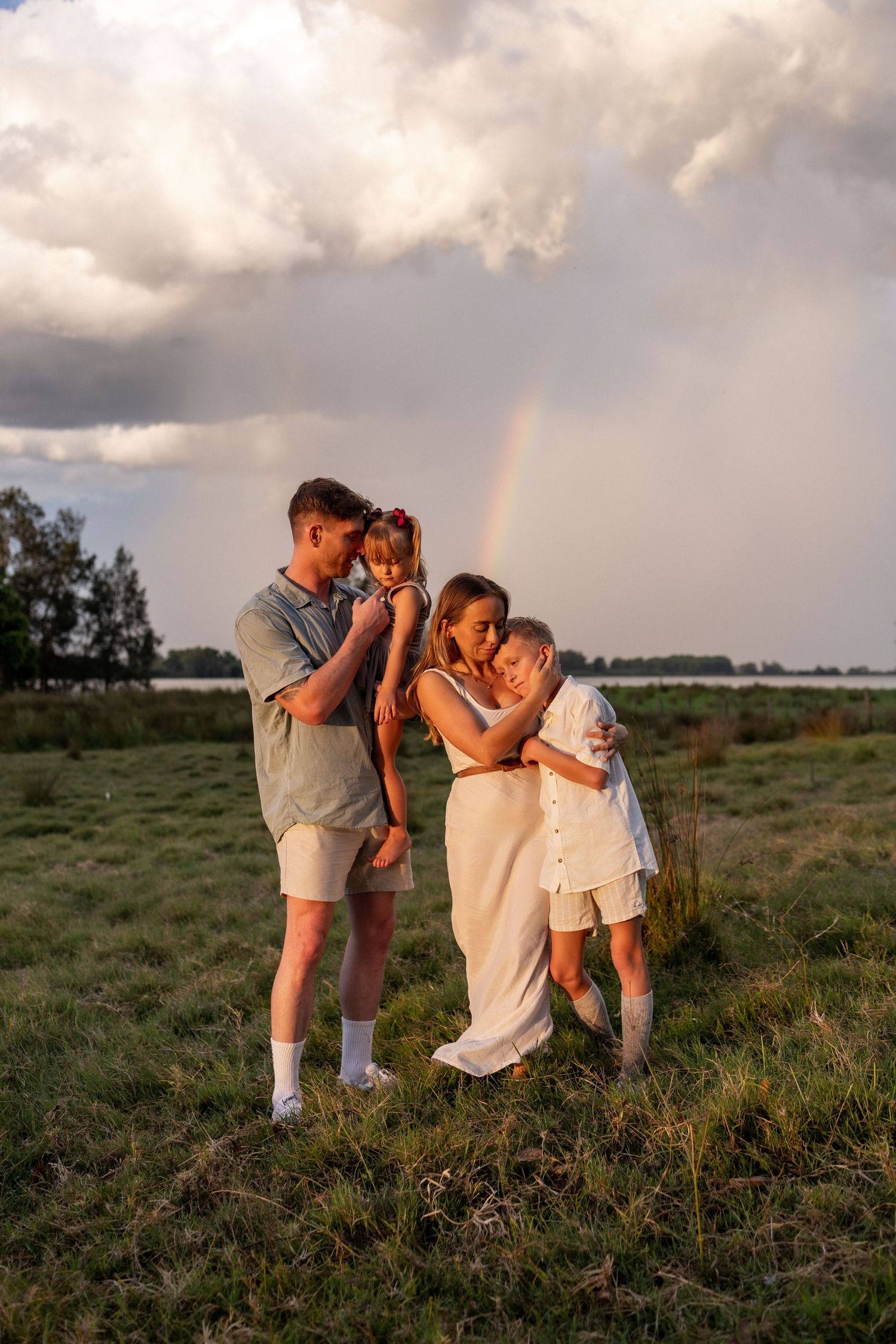 Family of four embracing in a grassy field at sunset with a rainbow and stormy clouds in the background.