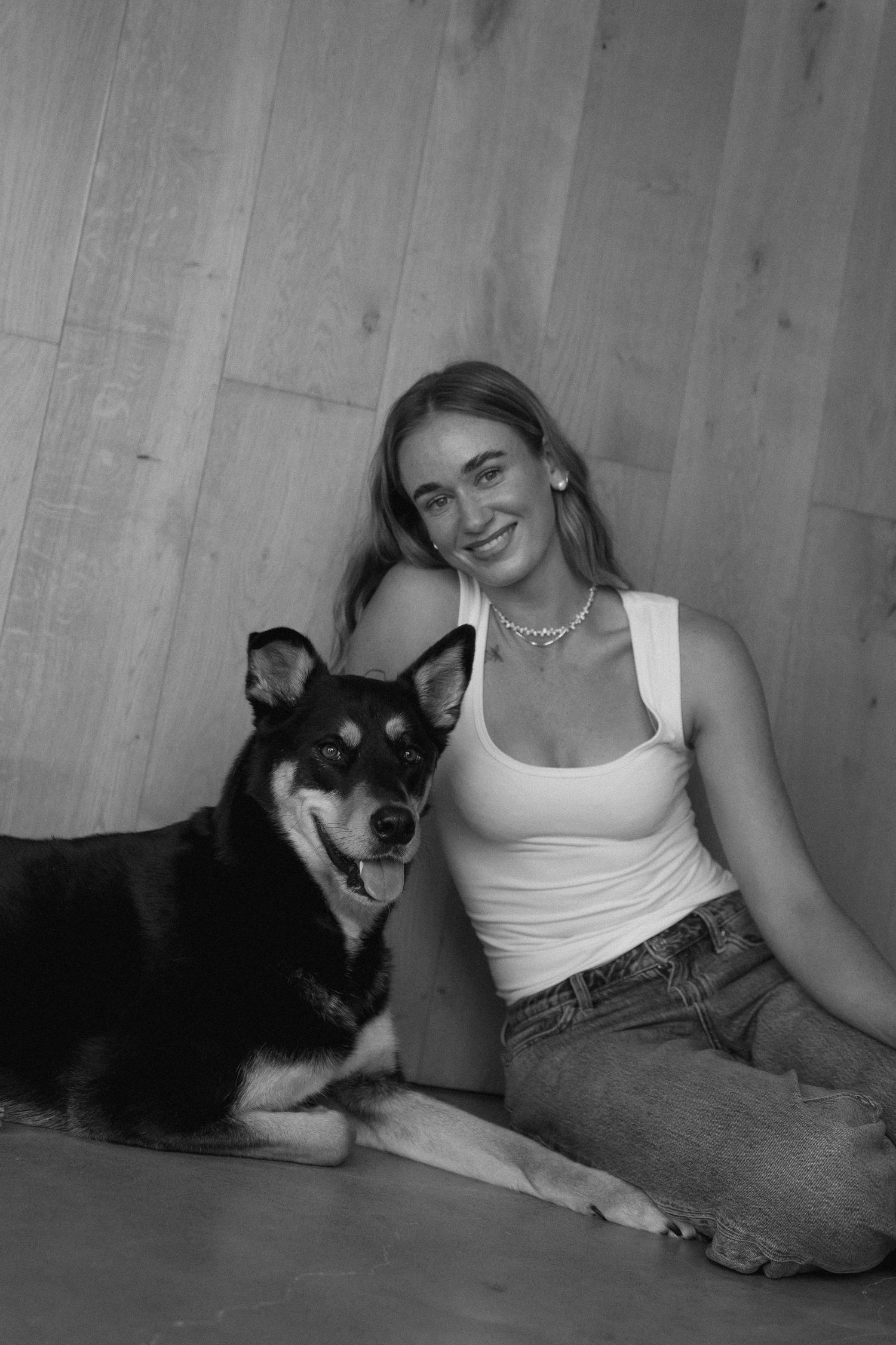 A smiling young woman in a white tank top and jeans sits on the floor beside a black and tan dog against a wood-paneled wall.